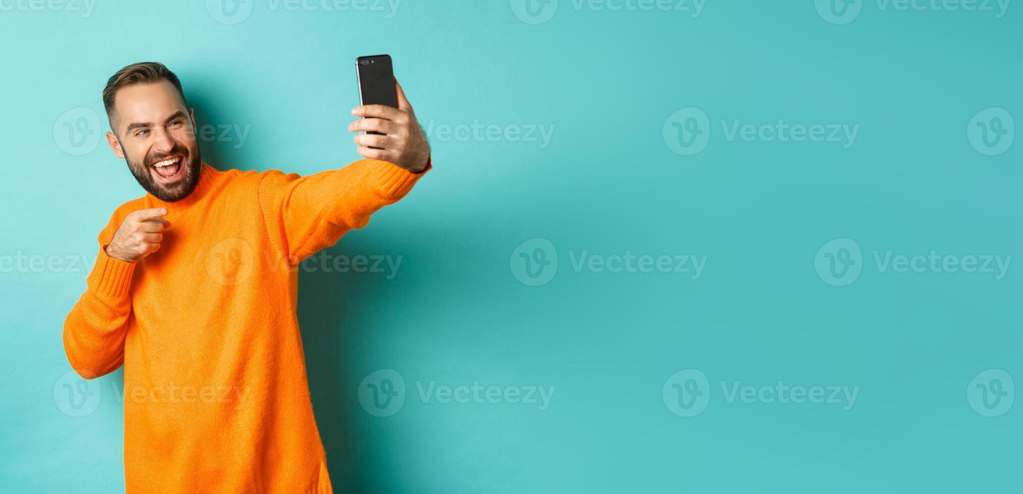 Handsome man calling on mobile phone, taking selfie and pointing at smartphone camera, standing over turquoise background photo