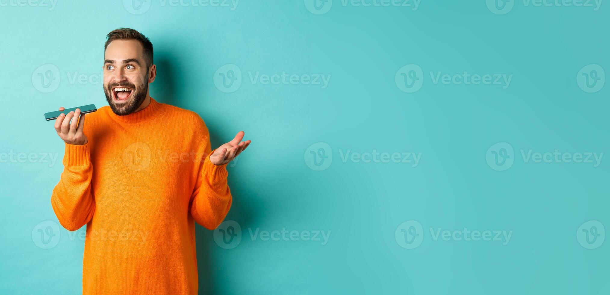 Happy man talking on speakerphone, gesturing and recording voice message on mobile phone, standing in orange sweater over light blue background photo