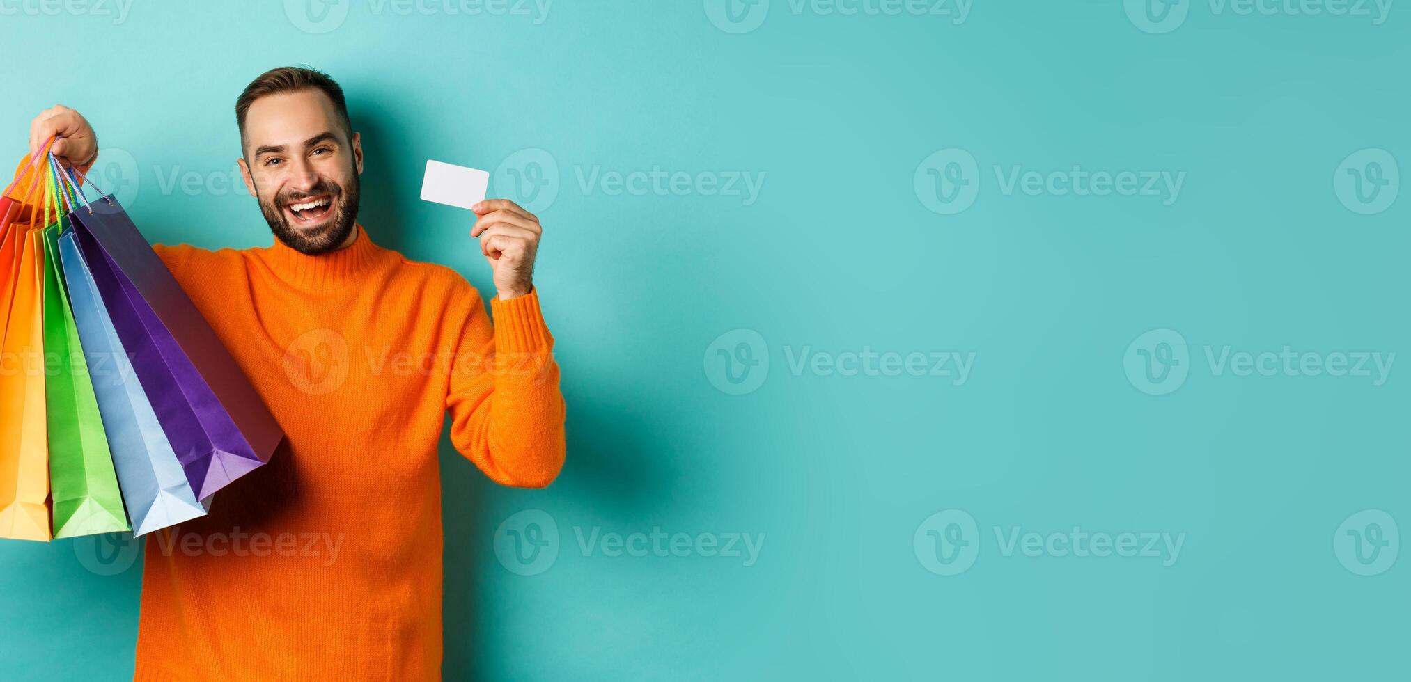 Happy aduly man showing credit card and shopping bags, standing against turquoise background photo
