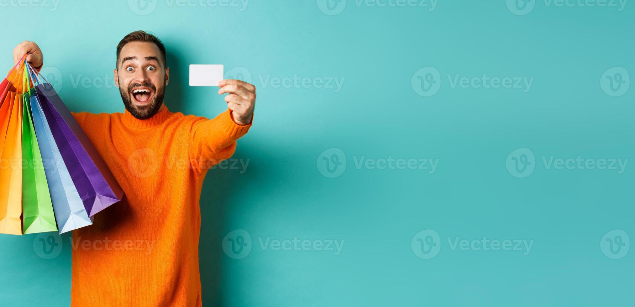 Happy aduly man showing credit card and shopping bags, standing against turquoise background photo
