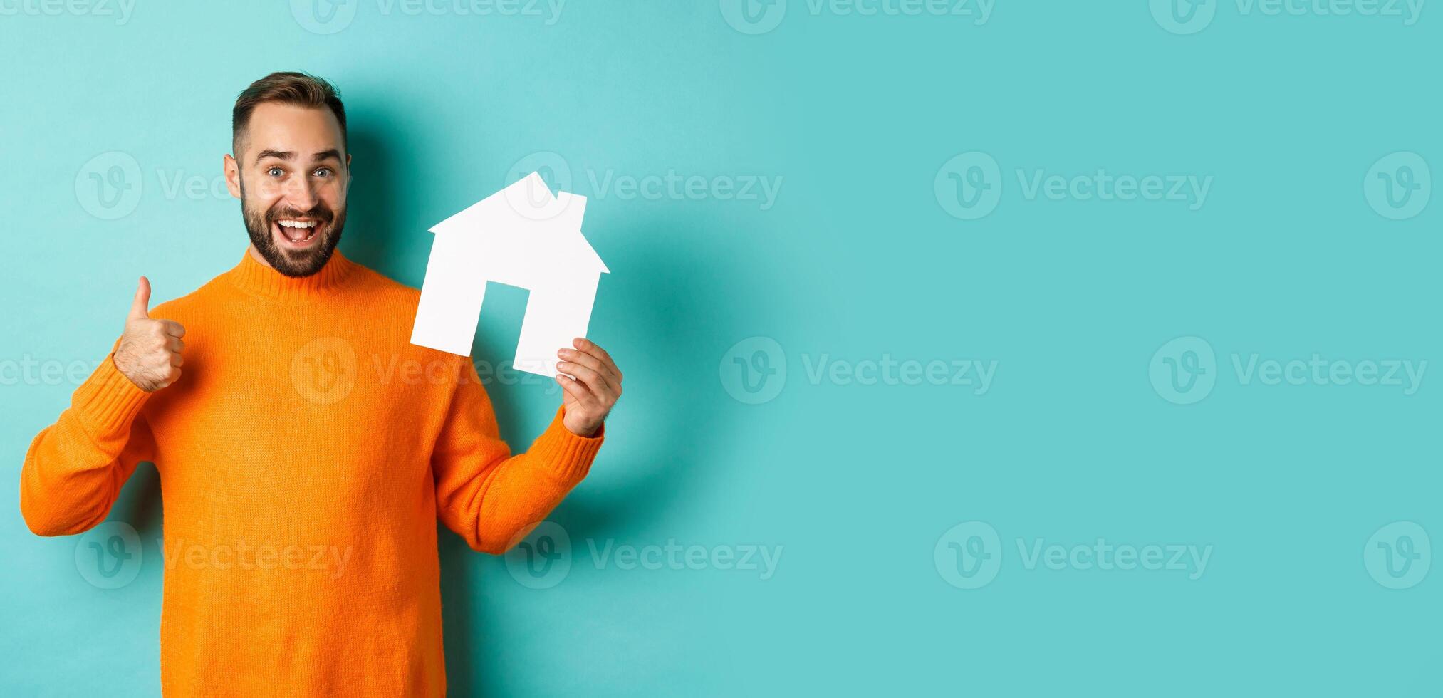 Real estate concept. Satisfied man recommending agency, showing thumb up and house maket, standing over blue background photo