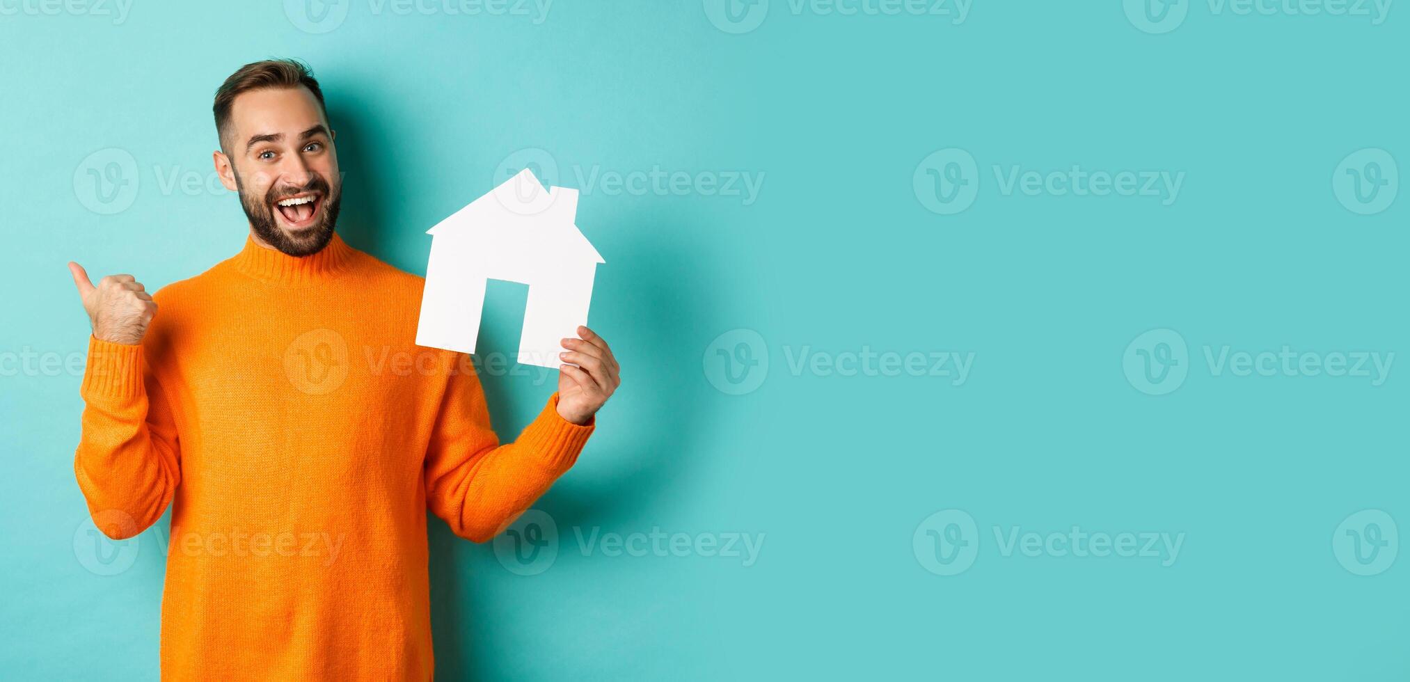 Real estate concept. Satisfied man recommending agency, pointing at copy space banner with thumb, showing paper house maket photo