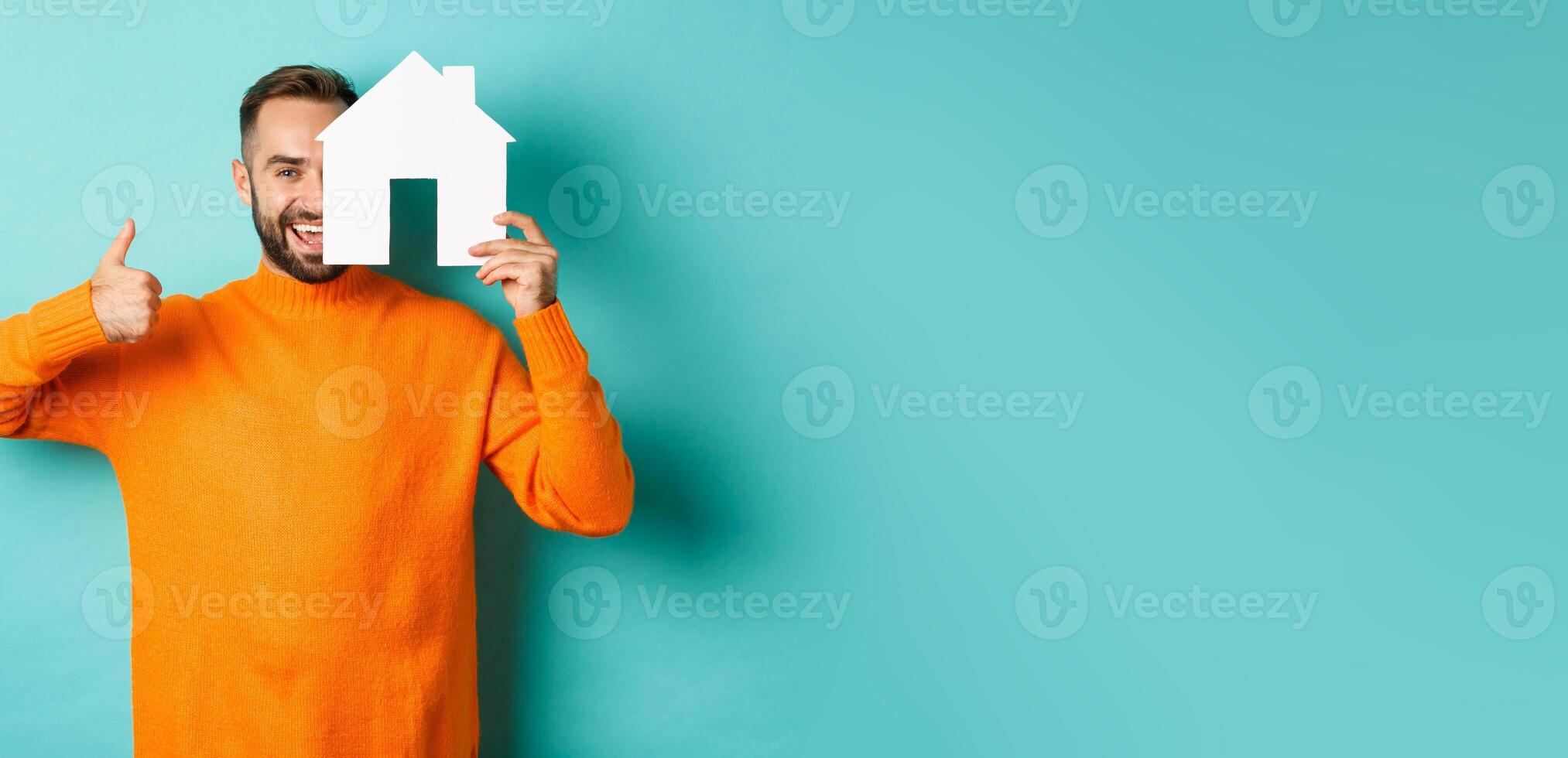 Real estate concept. Satisfied man recommending agency, showing thumb up and house maket, standing over blue background photo