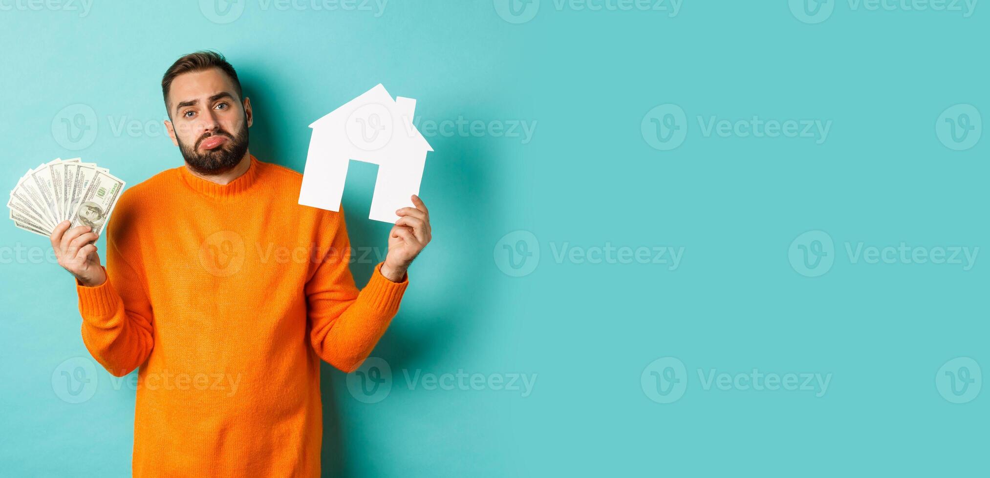 Real estate concept and mortgage concept. Sad and confused man showing money and paper house maket, shrugging upset, standing over light blue background photo