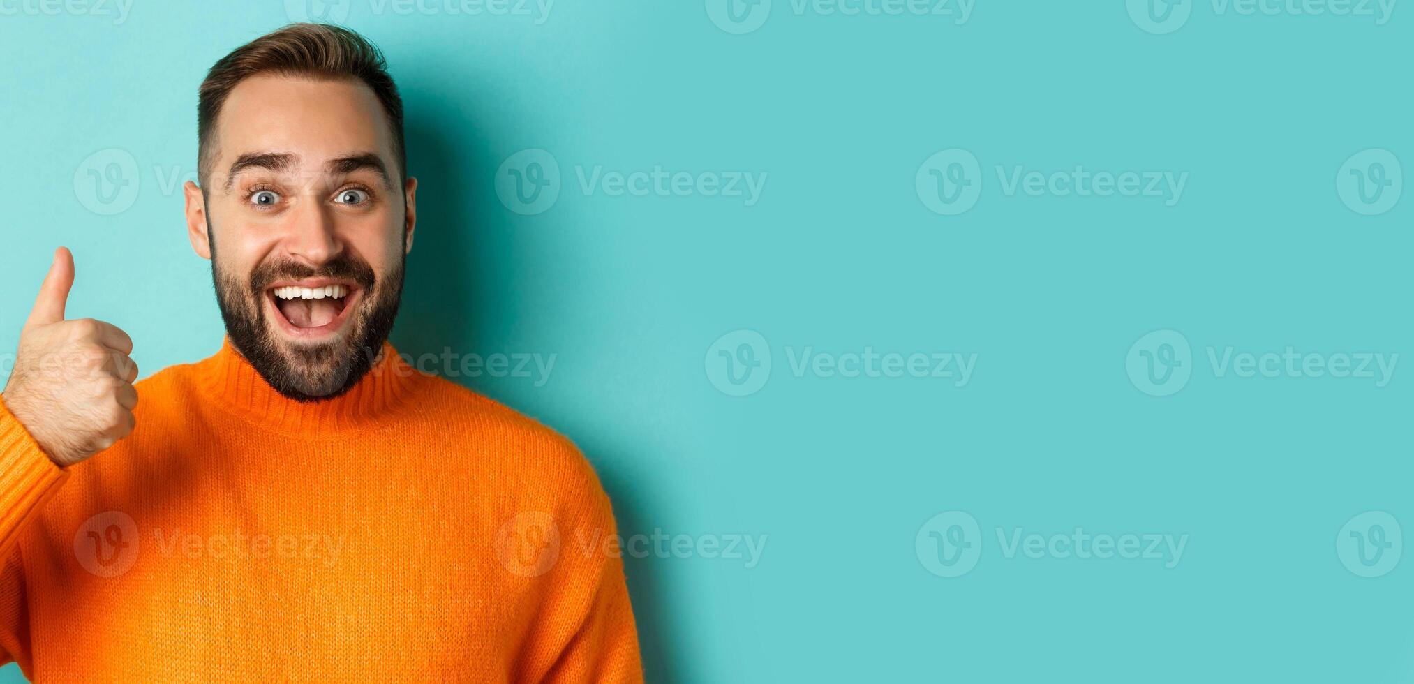 Close-up of cheerful male model showing thumb-up, smiling pleased, standing over light blue background photo