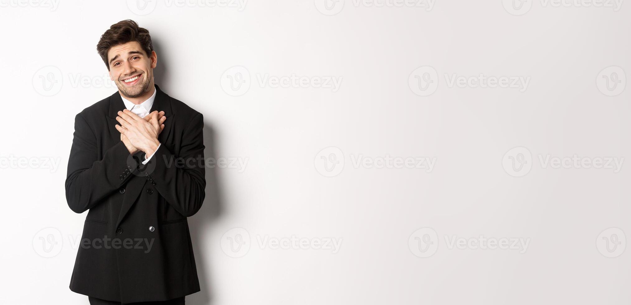 Portrait of happy and pleased handsome man in party suit, holding hands on heart and sighing flattered, saying thank you, standing grateful over white background photo