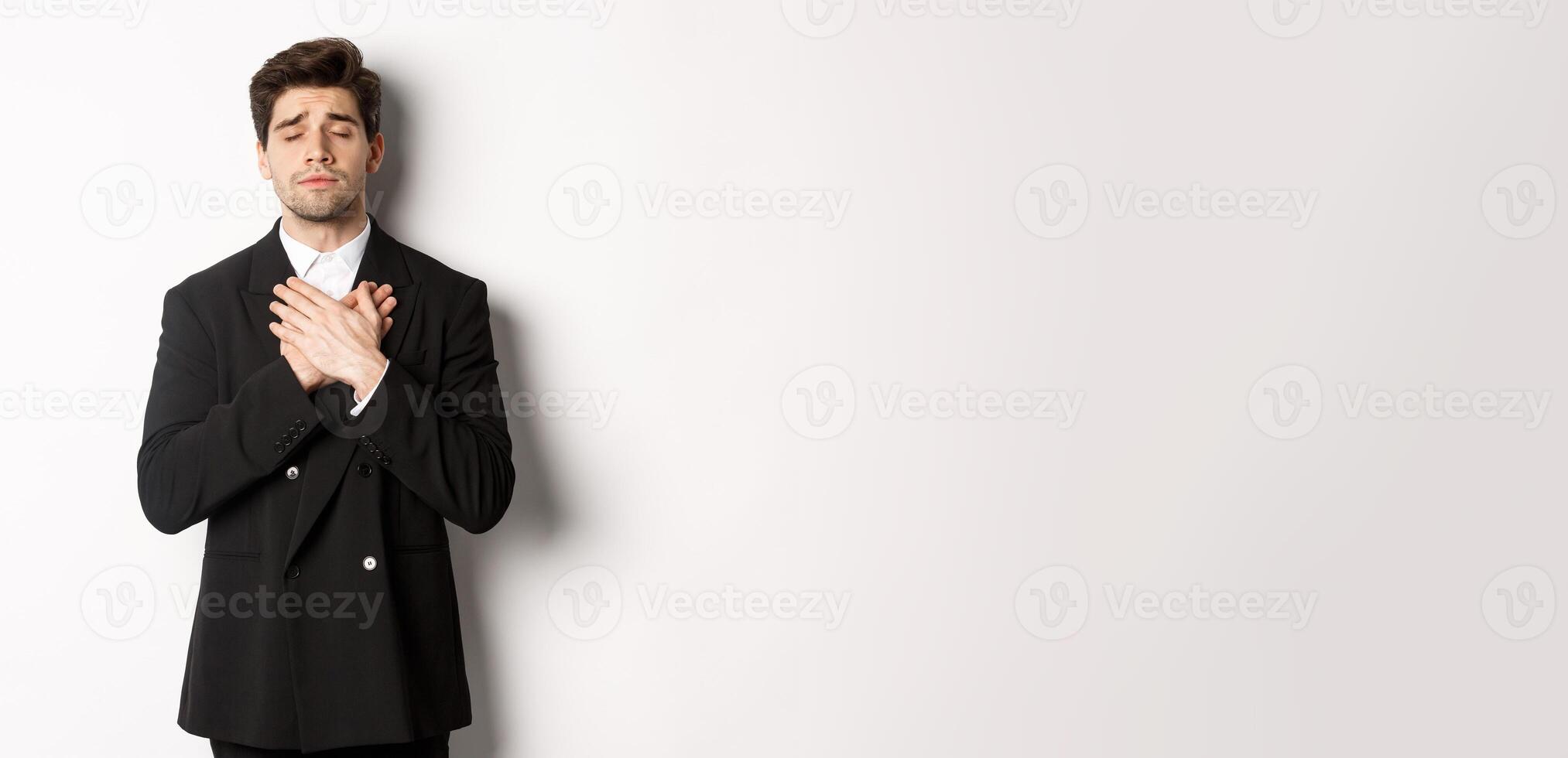 Portrait of dreamy and heartfelt bearded man in suit, close eyes and hold hands on heart, remember something, standing nostalgic over white background photo