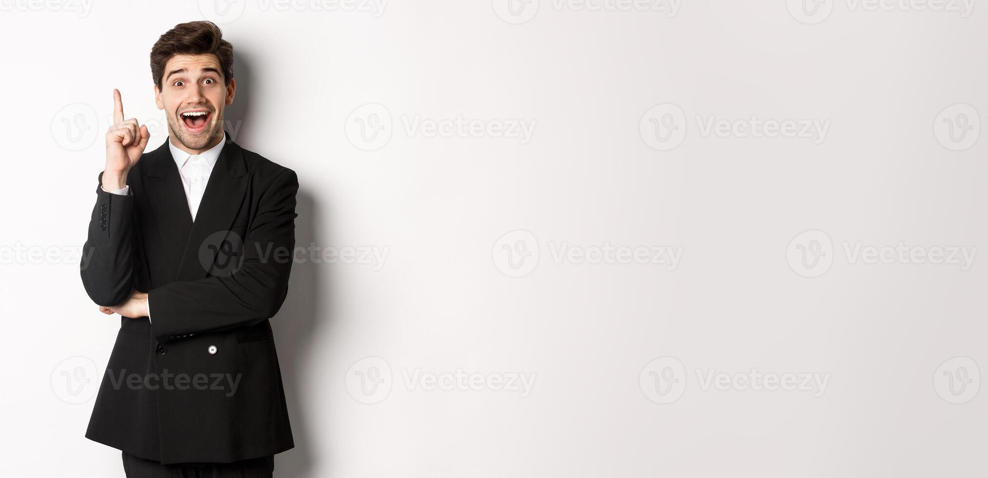 Portrait of attractive man in suit, having an idea, standing excited and raising one finger to tell suggestion, think-up solution, standing over white background photo