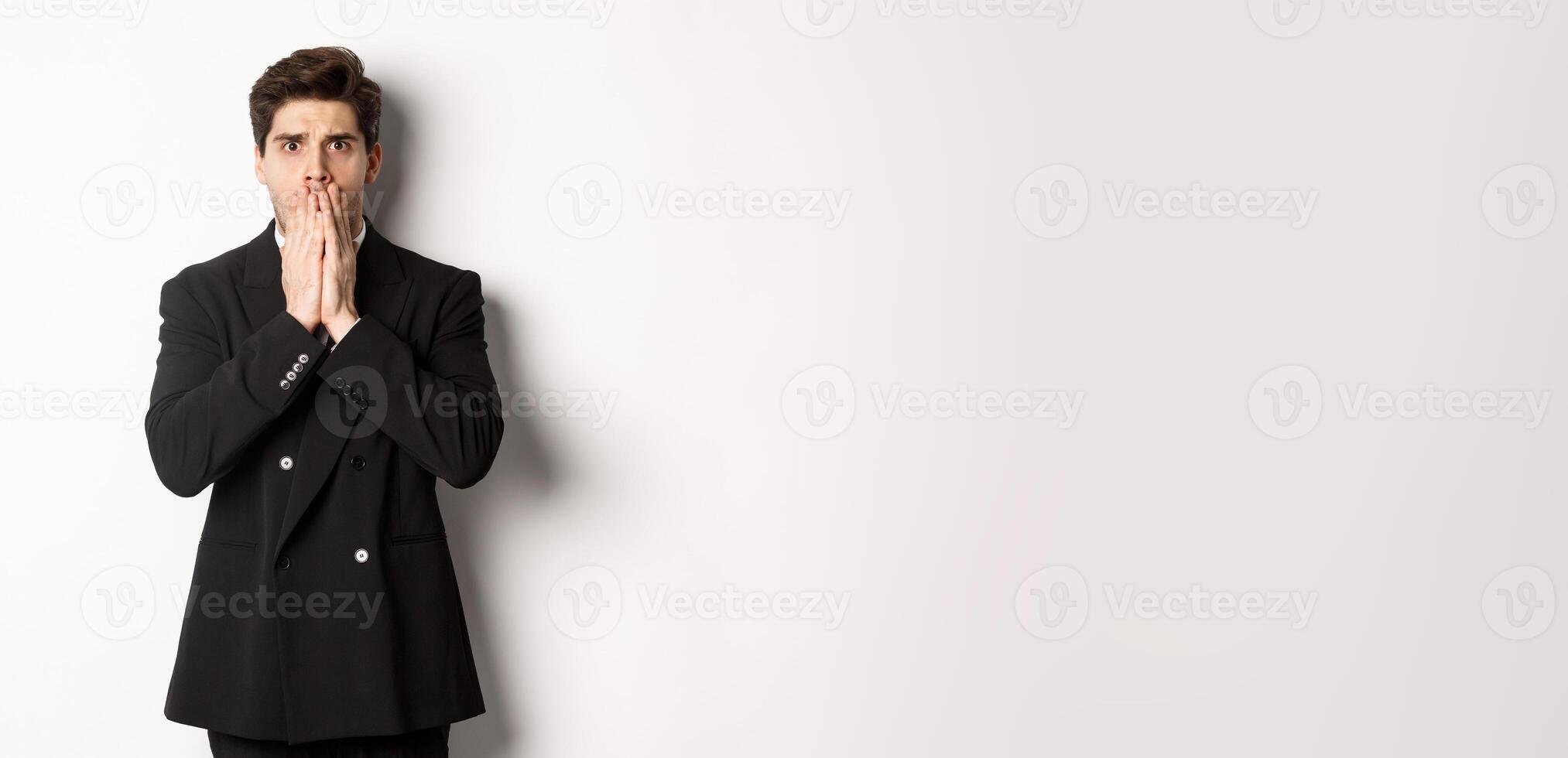 Portrait of shocked handsome businessman in suit, reacting to terrible situation, gasping and covering mouth with hands, standing startled against white background photo
