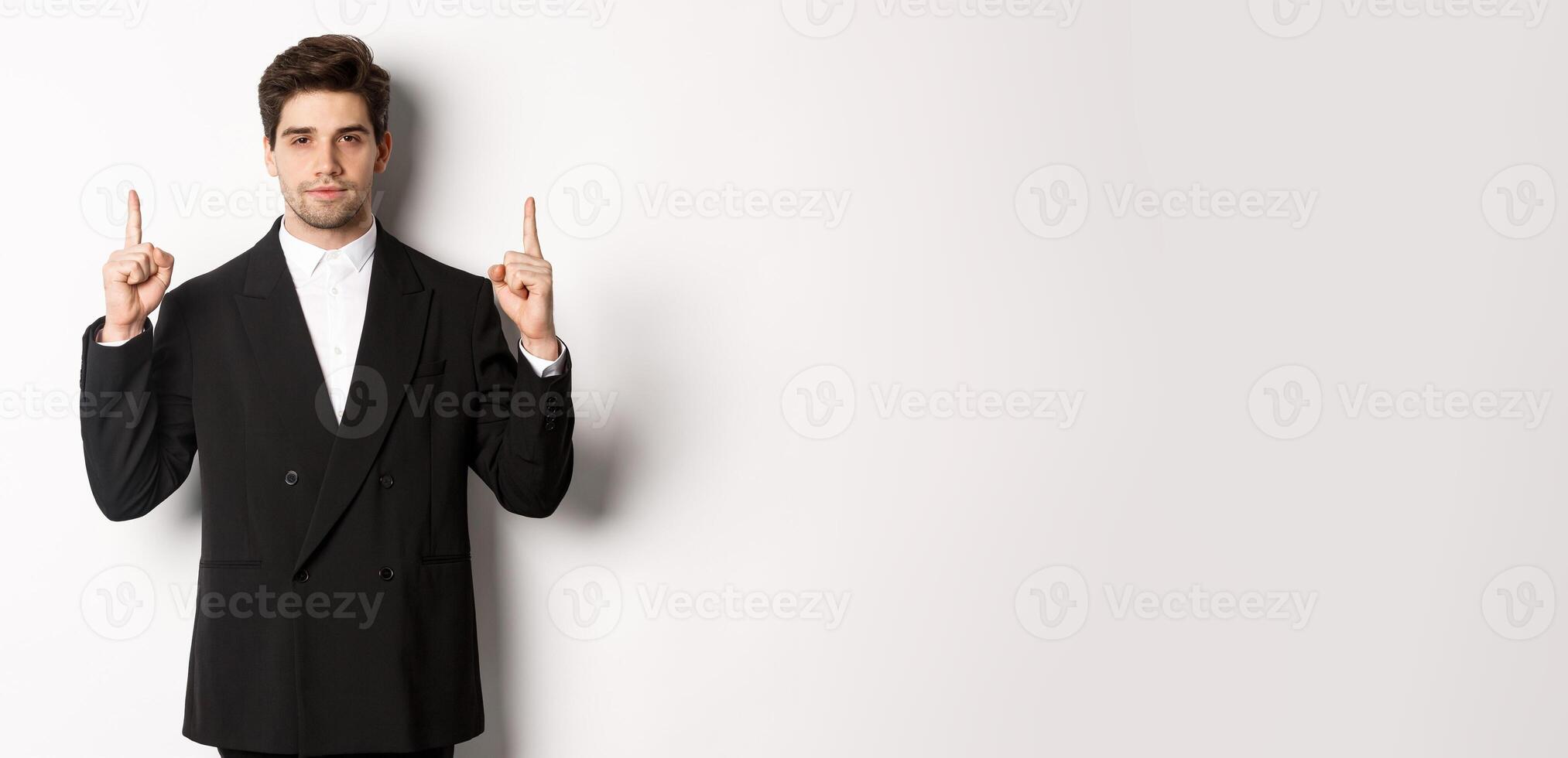 Image of confident and handsome man in formal suit, pointing fingers up, showing copy space on white background photo