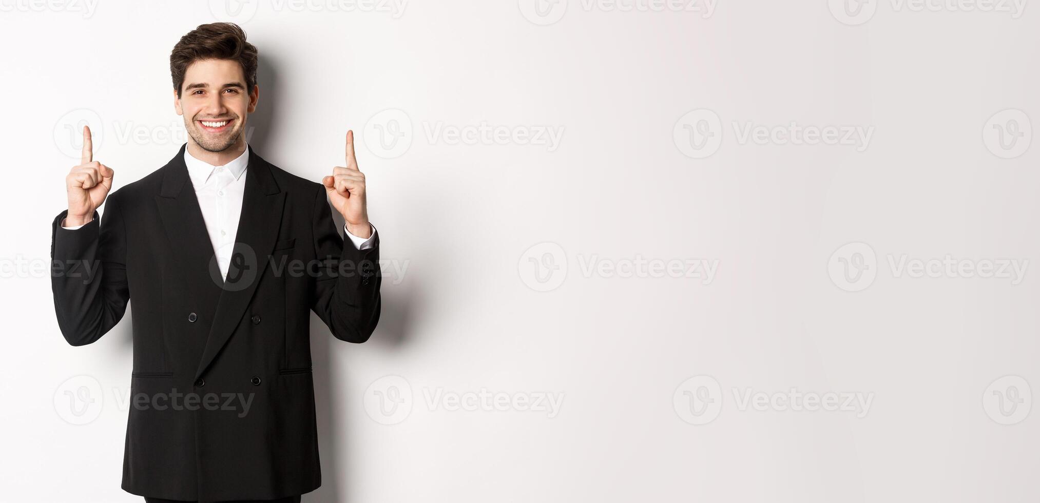 Portrait of attractive caucasian man in stylish black suit, pointing fingers up and smiling, showing christmas advertisement, standing over white background photo