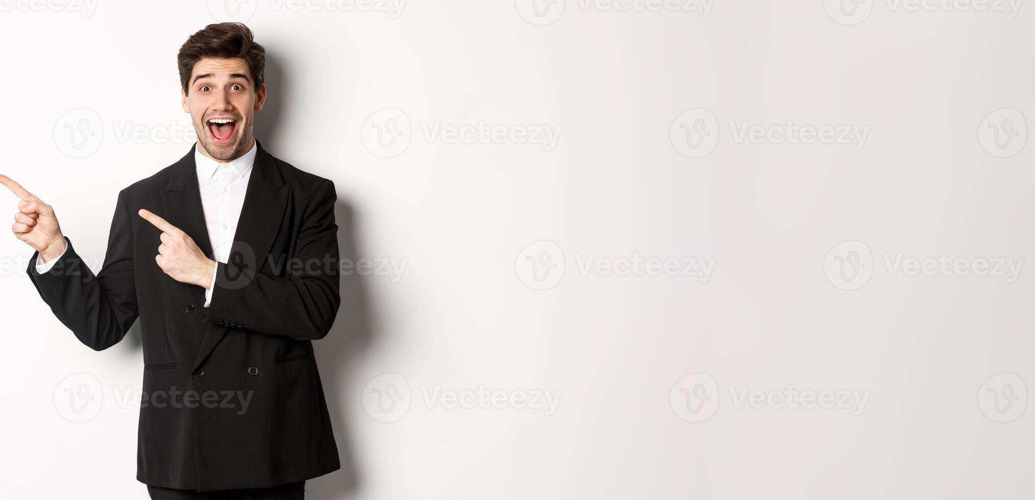Portrait of excited and happy man in suit showing advertisement, pointing fingers at upper left corner and smiling amazed, standing over white background photo
