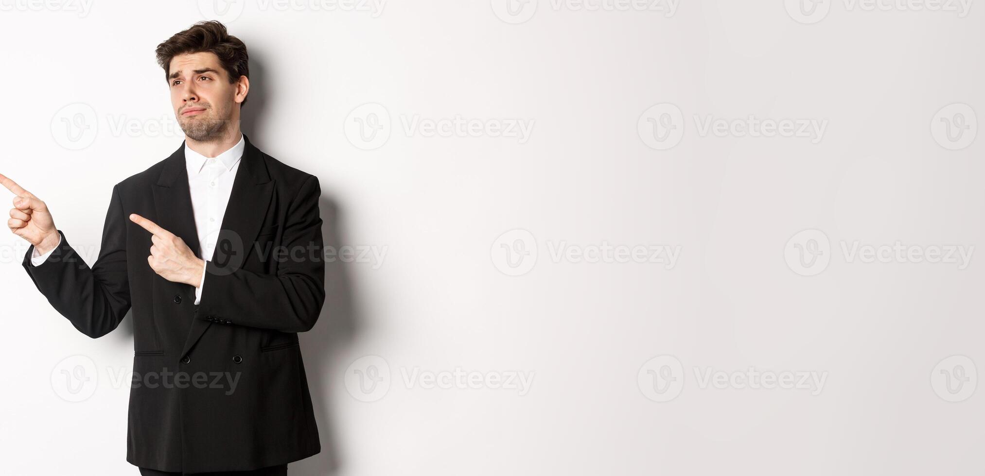 Image of upset and disappointed handsome guy in formal suit, pointing and looking left with sad face, standing over white background photo
