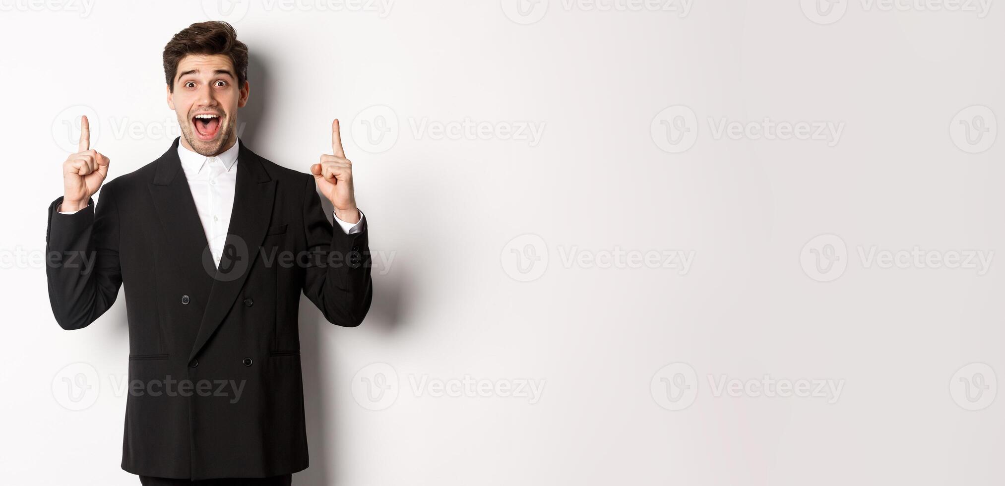 Image of handsome man in party suit, showing holidays promo, pointing fingers up and smiling amazed, standing over white background photo