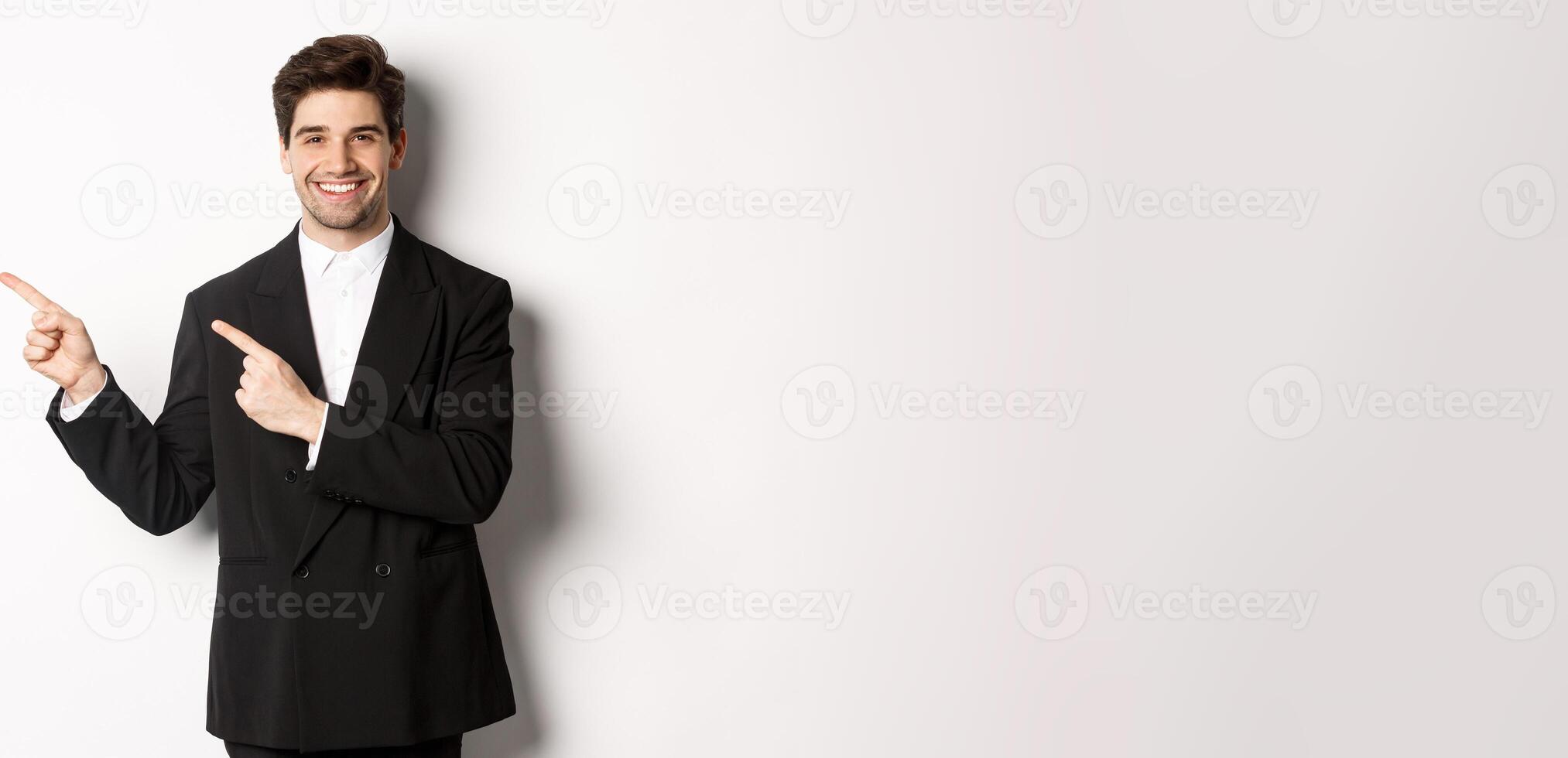 Portrait of successful, handsome businessman in suit, pointing fingers at upper left corner and smiling, showing advertisement, standing over white background photo