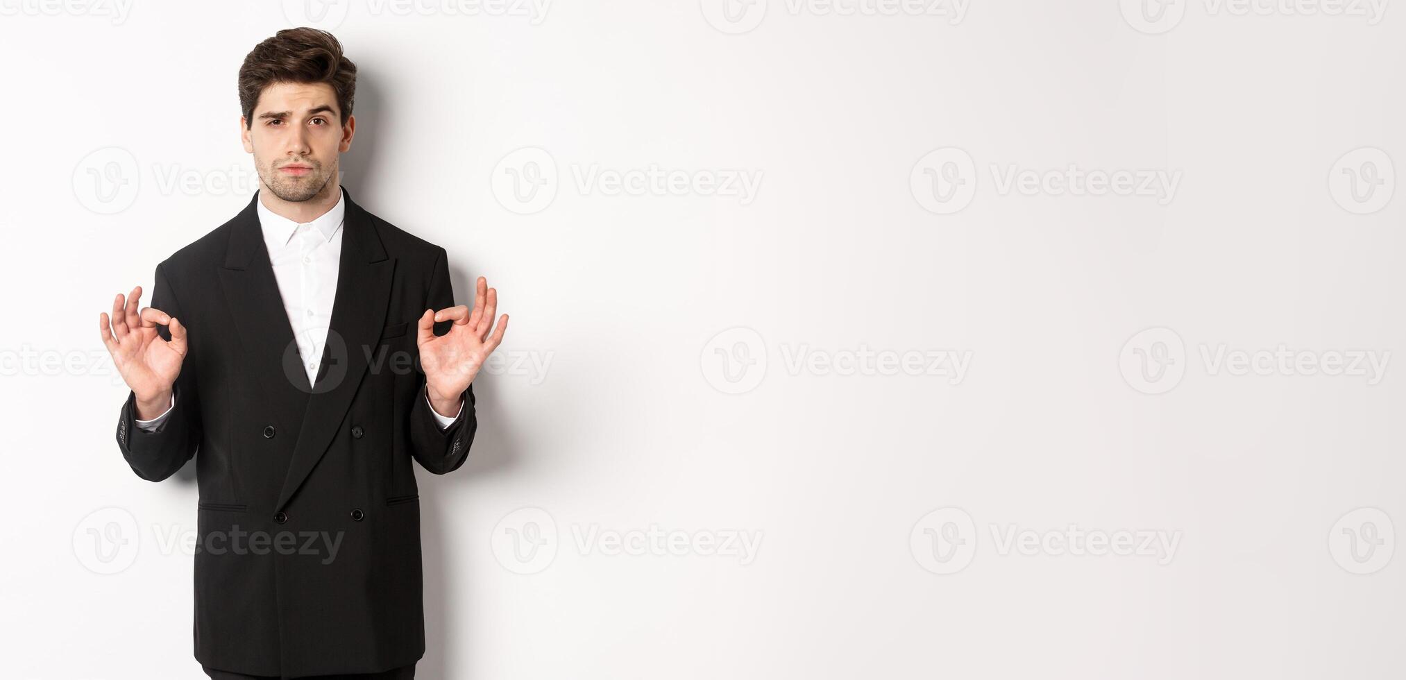 Concept of new year party, celebration and lifestyle. Portrait of confident good-looking man in black suit, showing okay sign and approve something, standing over white background photo