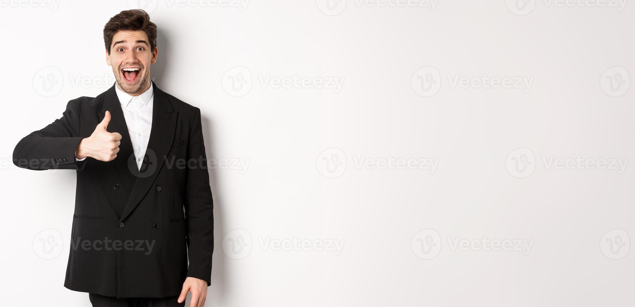 Portrait of amazed and impressed handsome man in black suit, showing thumb-up, praise awesome product, liking something good, standing against white background photo