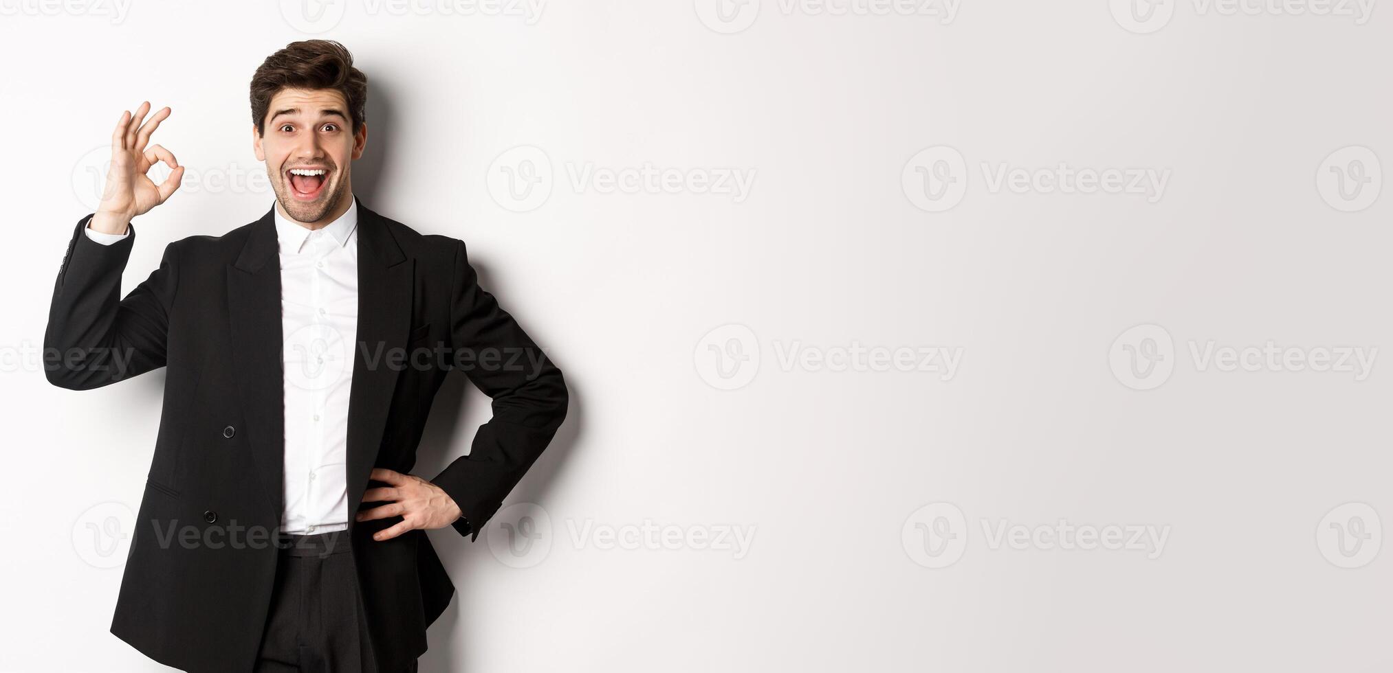 Concept of new year party, celebration and lifestyle. Portrait of pleased handsome guy in black suit, praise something good, showing okay sign in approval, standing over white background photo
