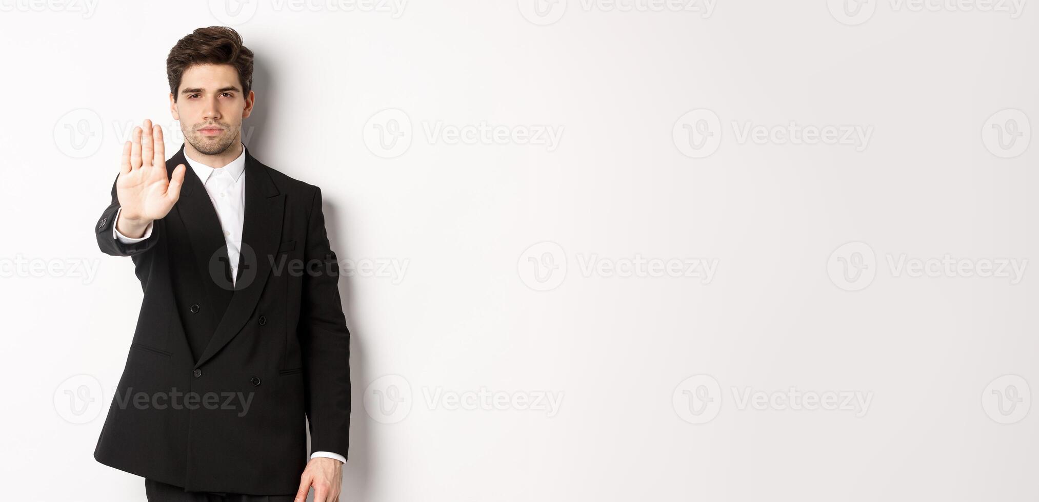 Portrait of serious handsome man in formal suit, extending hand to stop you, prohibit action, forbid and disagree with something, standing against white background photo