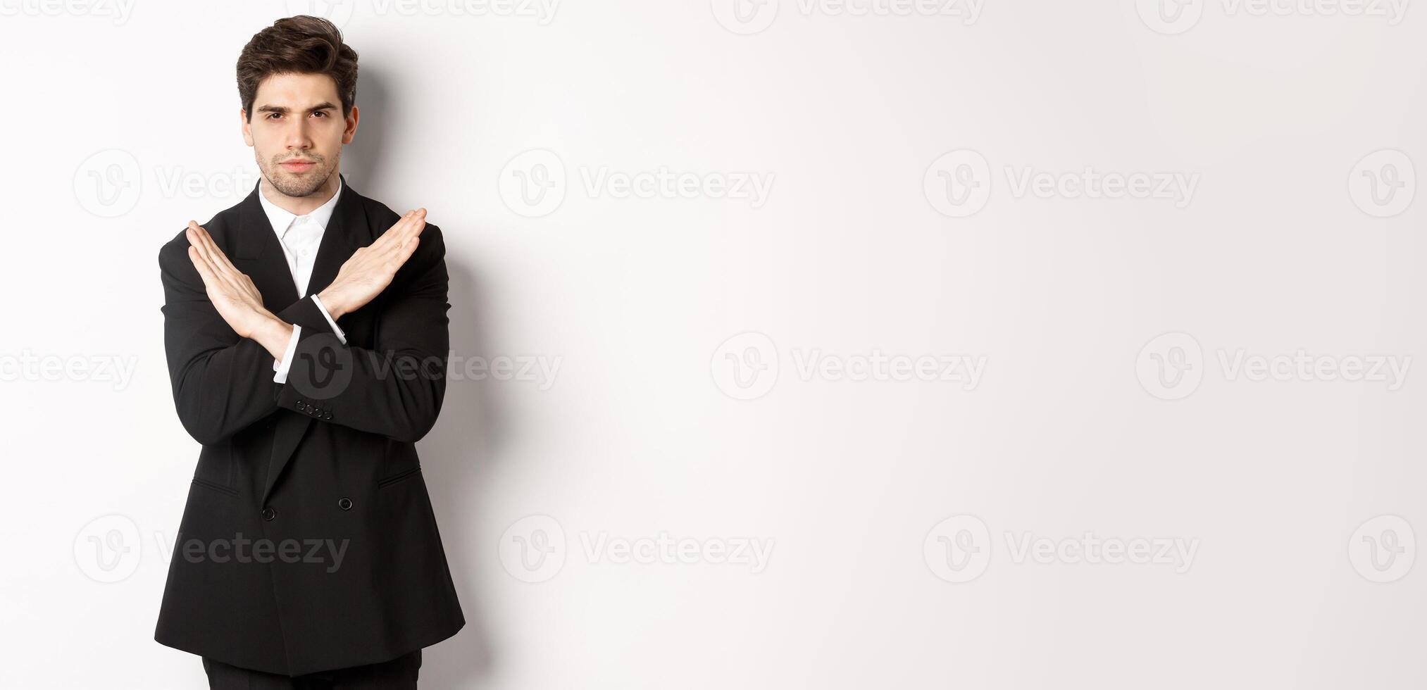 Portrait of serious and concerned man in black suit, showing stop gesture and frowning, making cross to prohibit or decline something, standing over white background photo