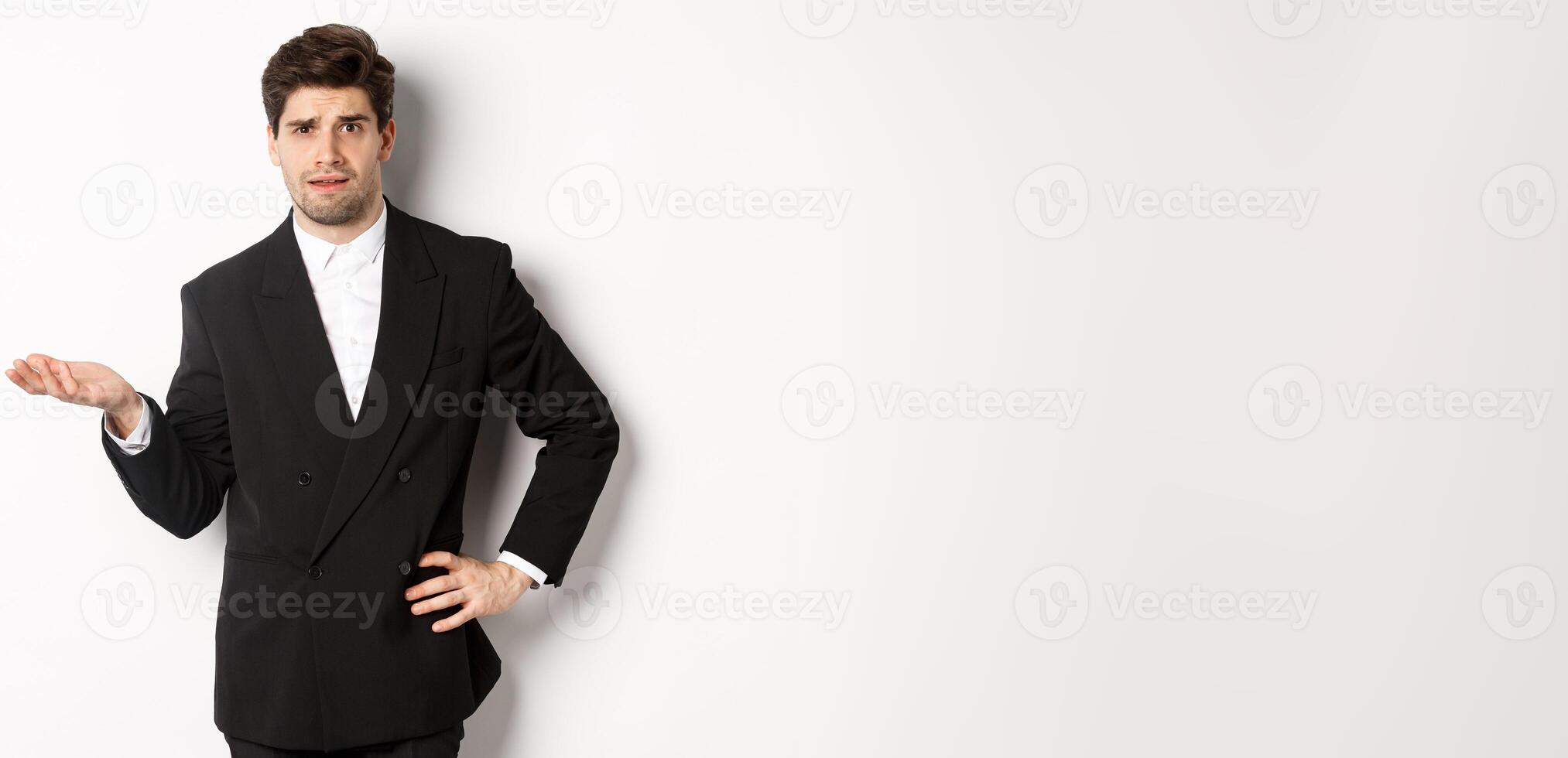 Image of confused handsome man in formal suit, raising hand and shrugging, cant understand something, standing against white background photo