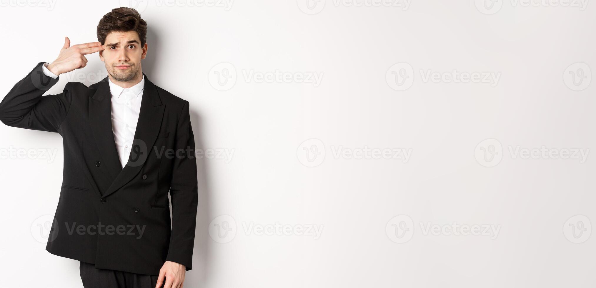 Portrait of distressed and troubled businessman in suit, pointing finger gun at head and looking bothered, shooting himself, standing against white background photo