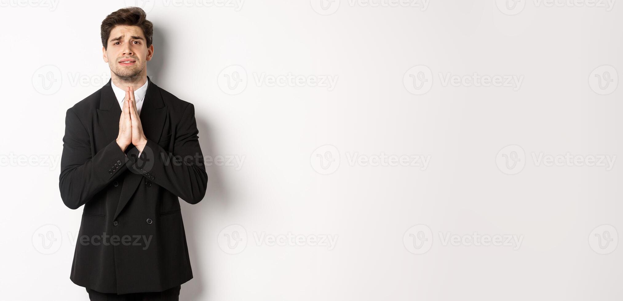 Image of handsome businessman in suit need help, begging you, holding hands in plead gesture and looking desperate, standing over white background photo