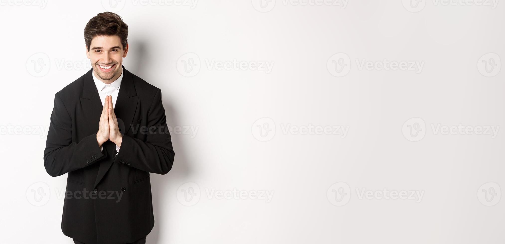 Portrait of handsome man in black suit, being grateful, saying thank you and bowing politely, smiling as holding hands together, expressing gratitude, standing over white background photo