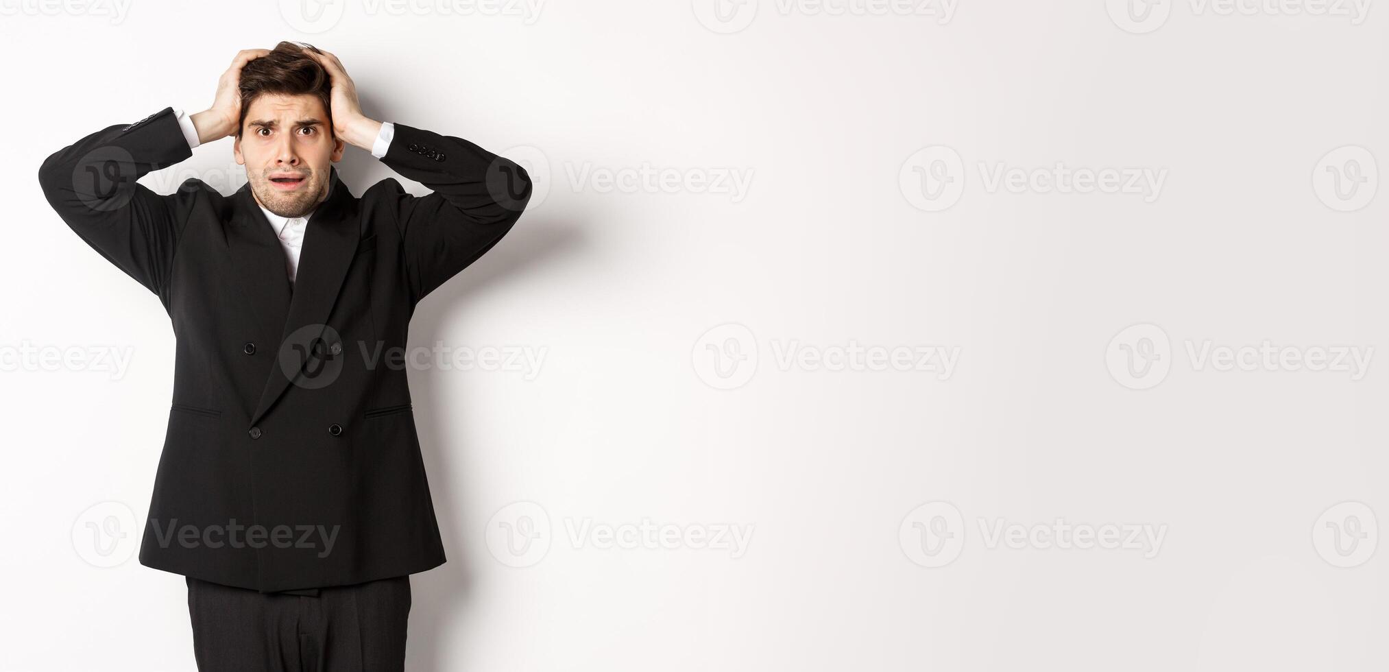 Frustrated and worried businessman in black suit, panicking as looking at trouble, holding hands on head alarmed, standing against white background photo