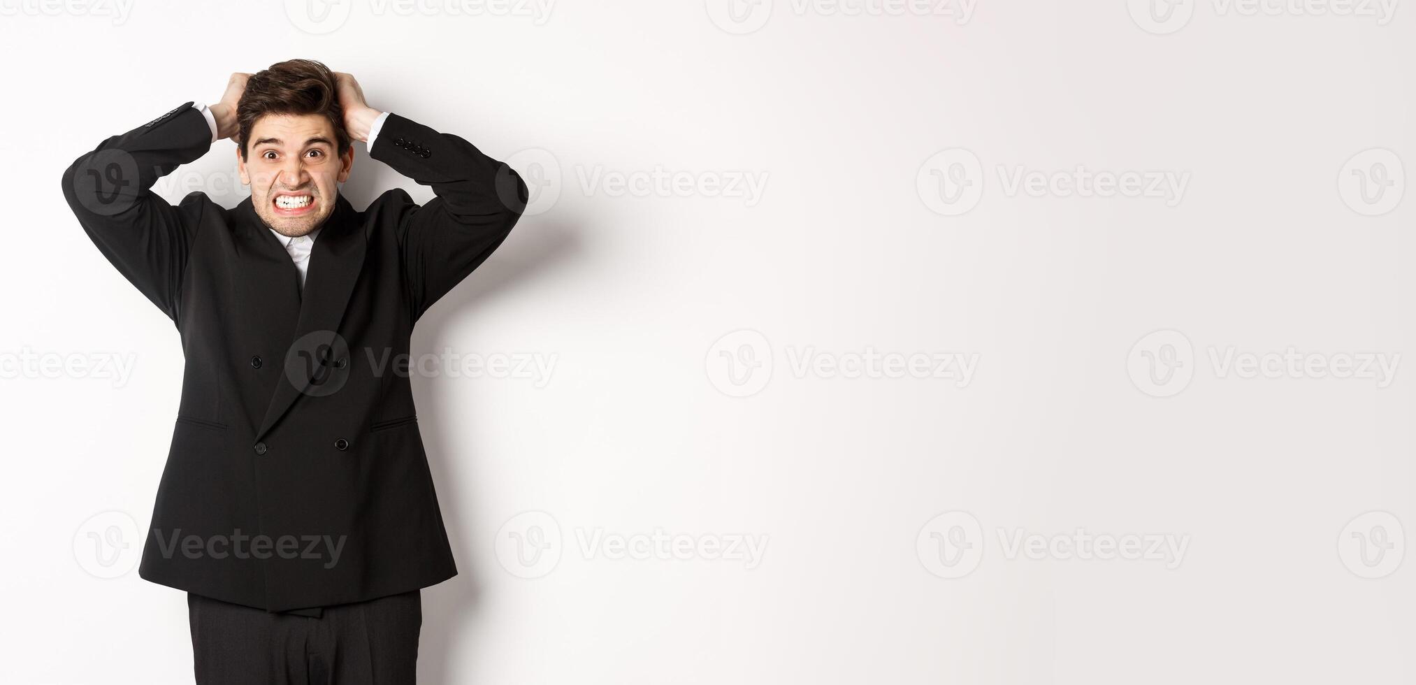 Image of frustrated and angry businessman in black suit, ripping hair on head and grimacing mad, standing tensed against white background photo