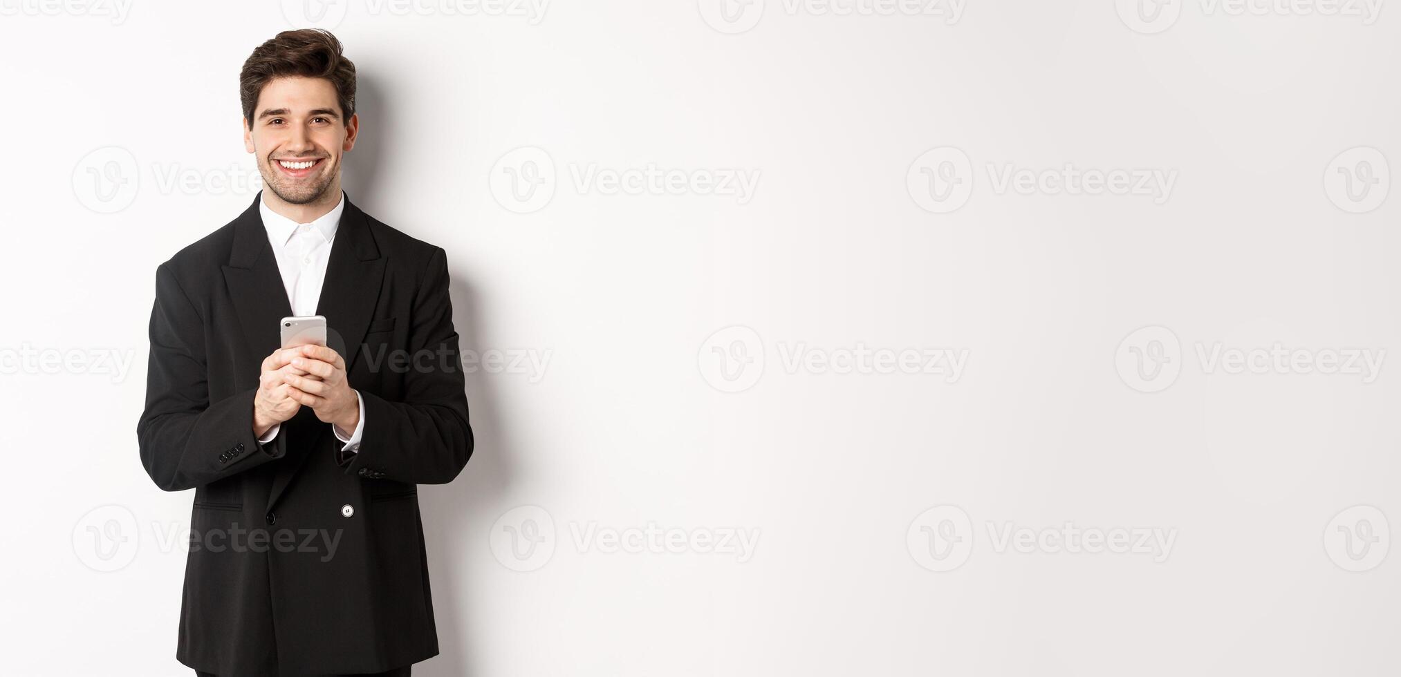 Portrait of successful, handsome businessman in suit, writing message on smartphone and smiling, standing over white background photo