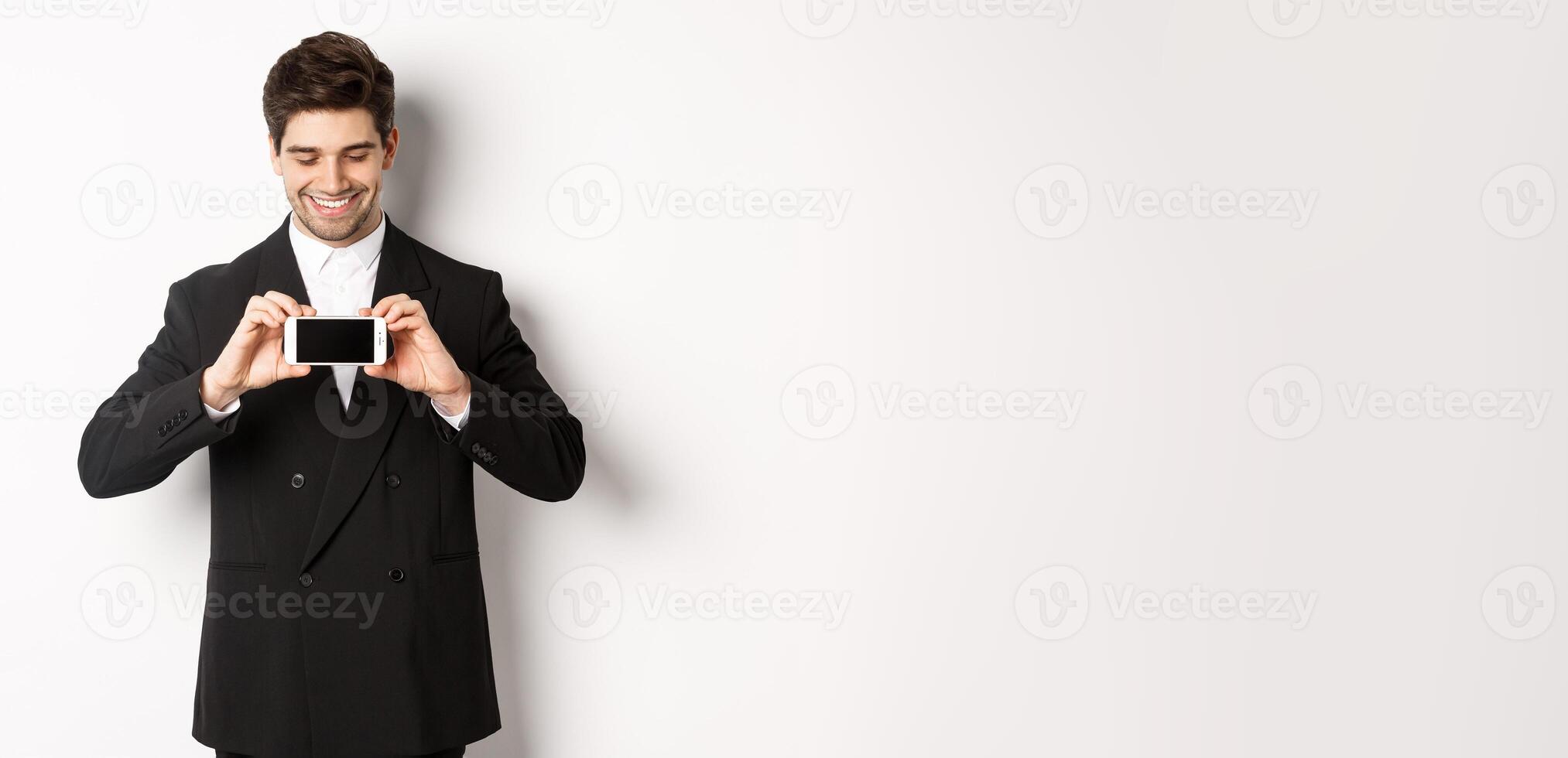 Portrait of handsome bearded man in black suit, showing you smartphone screen and smiling pleased, standing against white background photo
