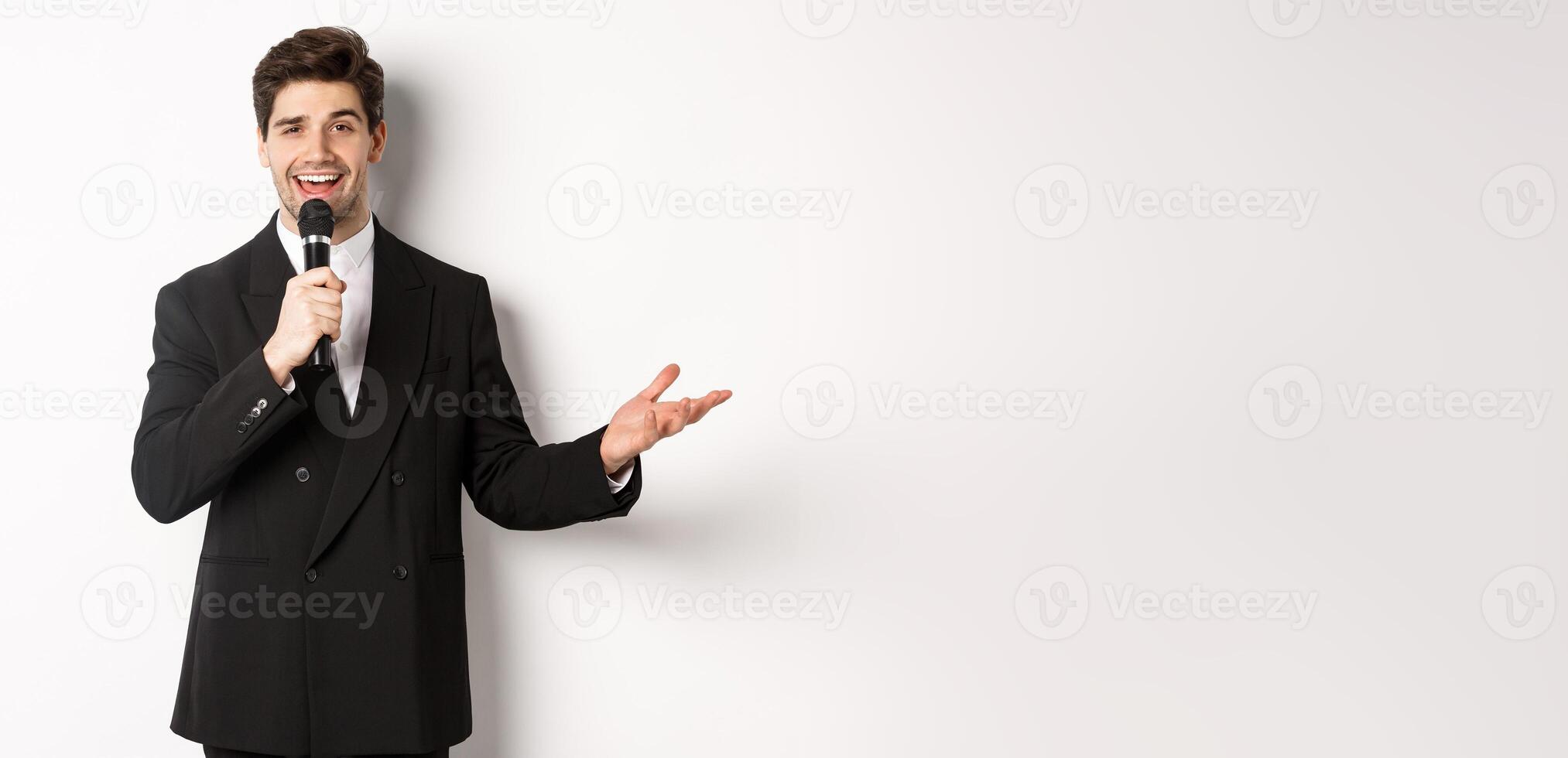 Portrait of handsome man in black suit singing a song, holding microphone and giving speech, standing against white background photo