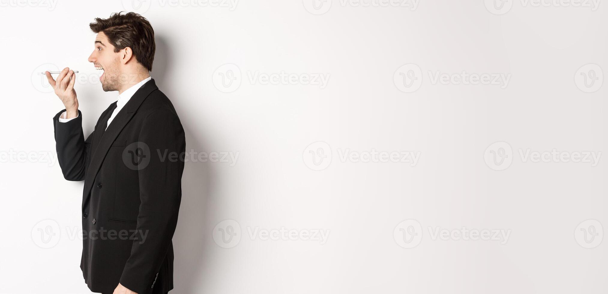 Profile shot of handsome businessman in black suit talking on speakerphone, smiling and looking happy, recording voice message, standing over white background photo
