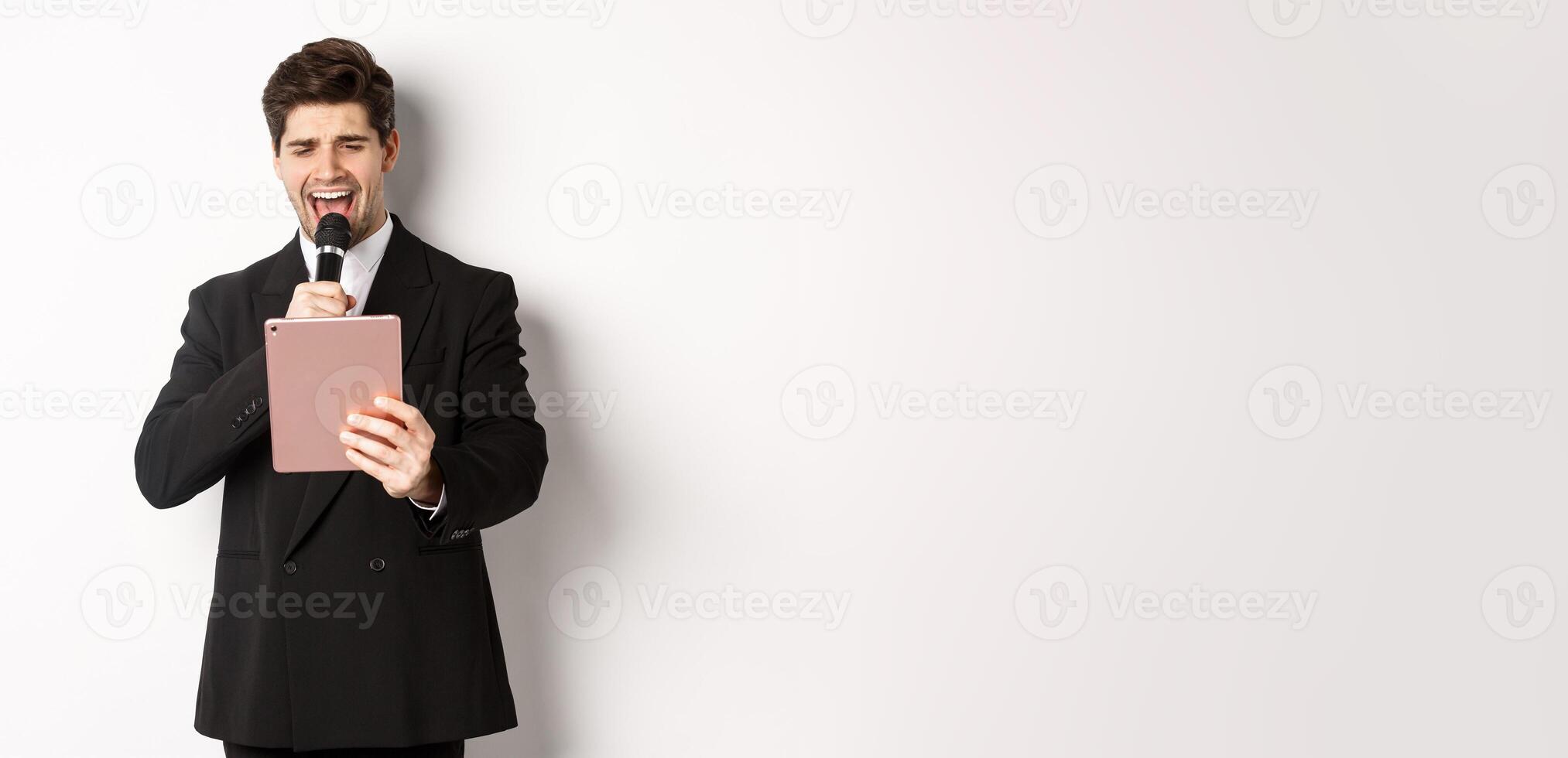 Image of handsome man in black suit, singing karaoke on digital tablet, holding microphone, standing over white background photo