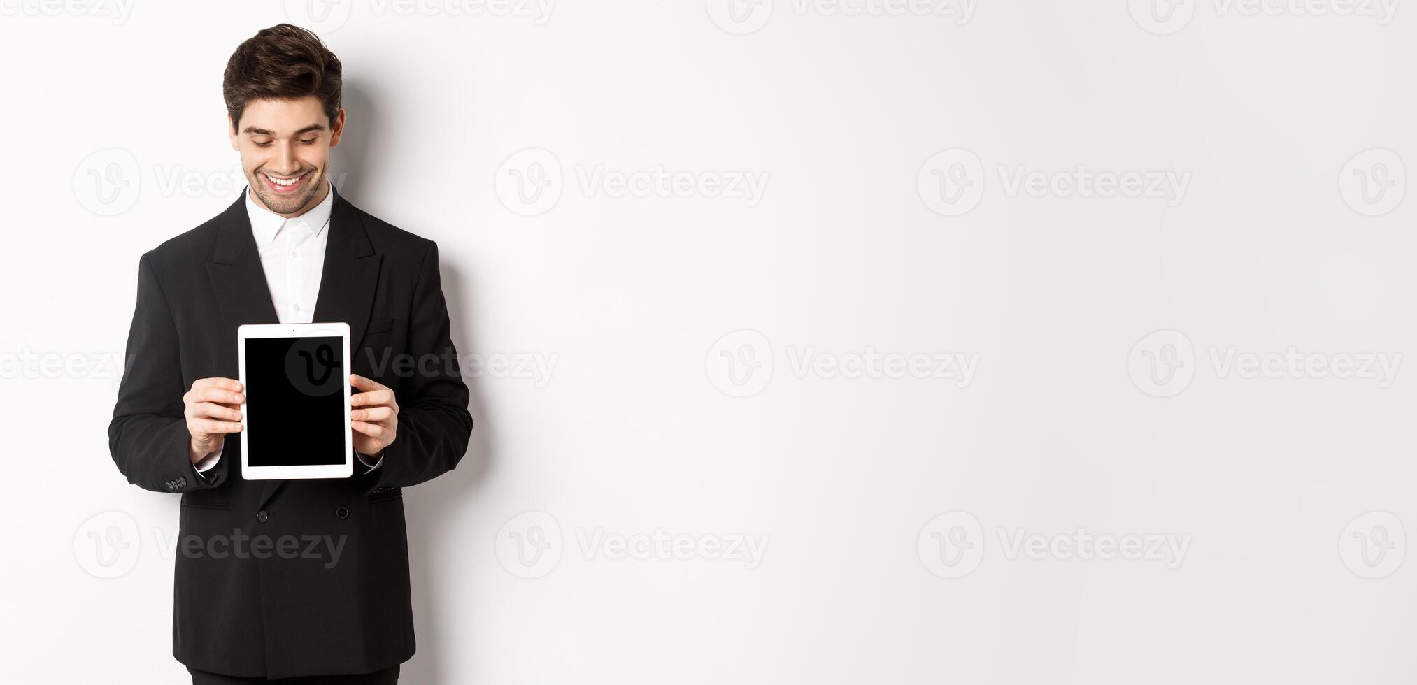 Image of good-looking male entrepreneur in black suit, looking down at digital tablet screen and showing advertisement, standing against white background photo