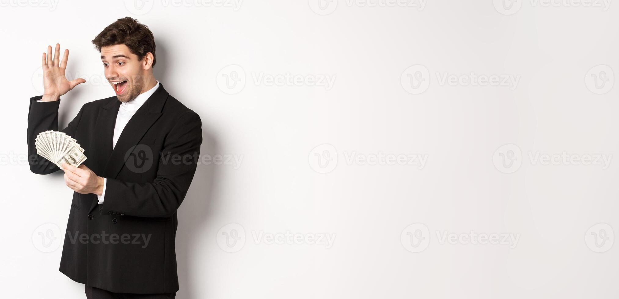 Image of successful businessman in black suit, looking at money and triumphing, earn cash, standing over white background photo