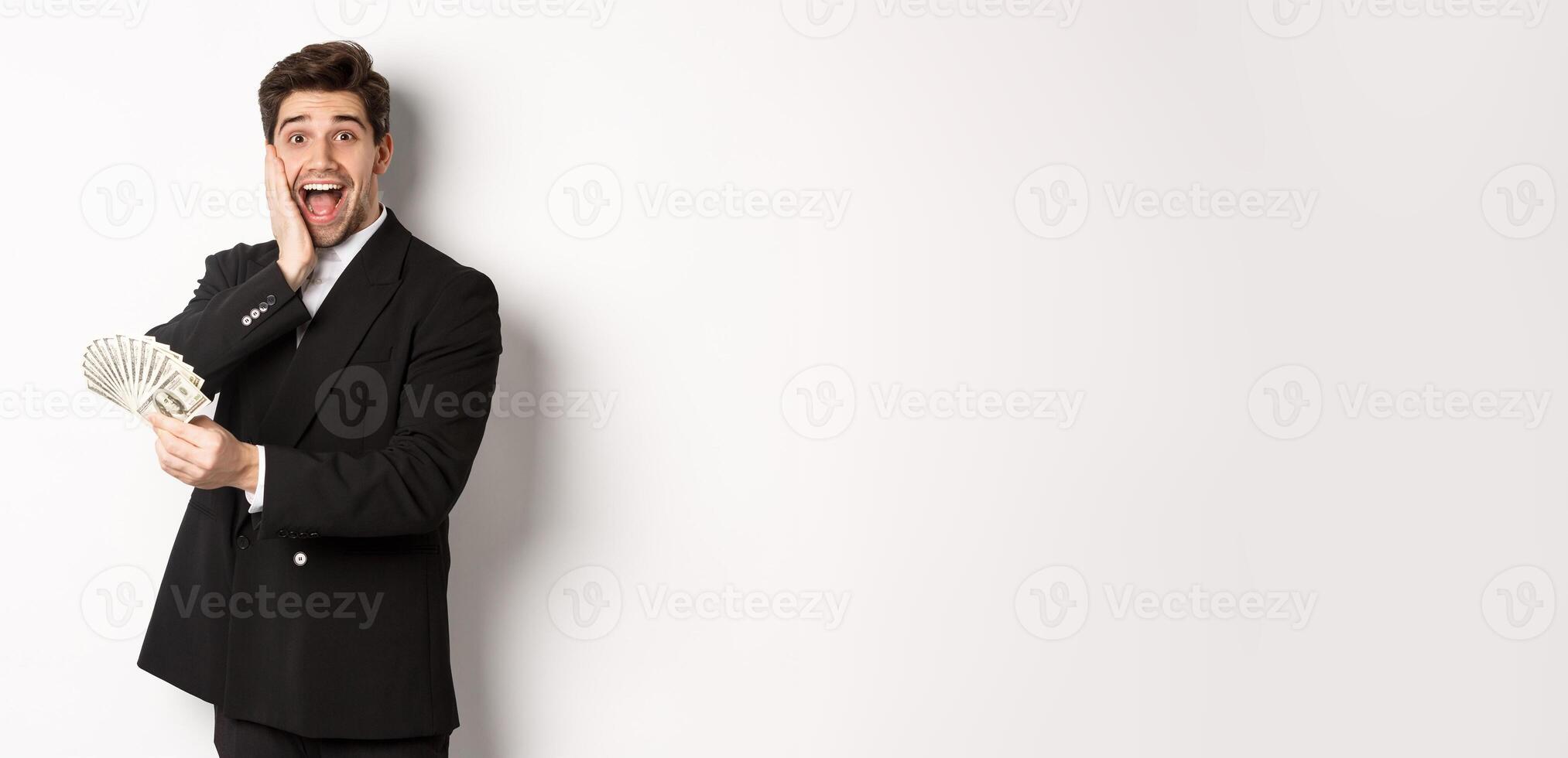 Image of rich and happy man in black suit, winning prize, holding money and looking excited at camera, standing over white background photo