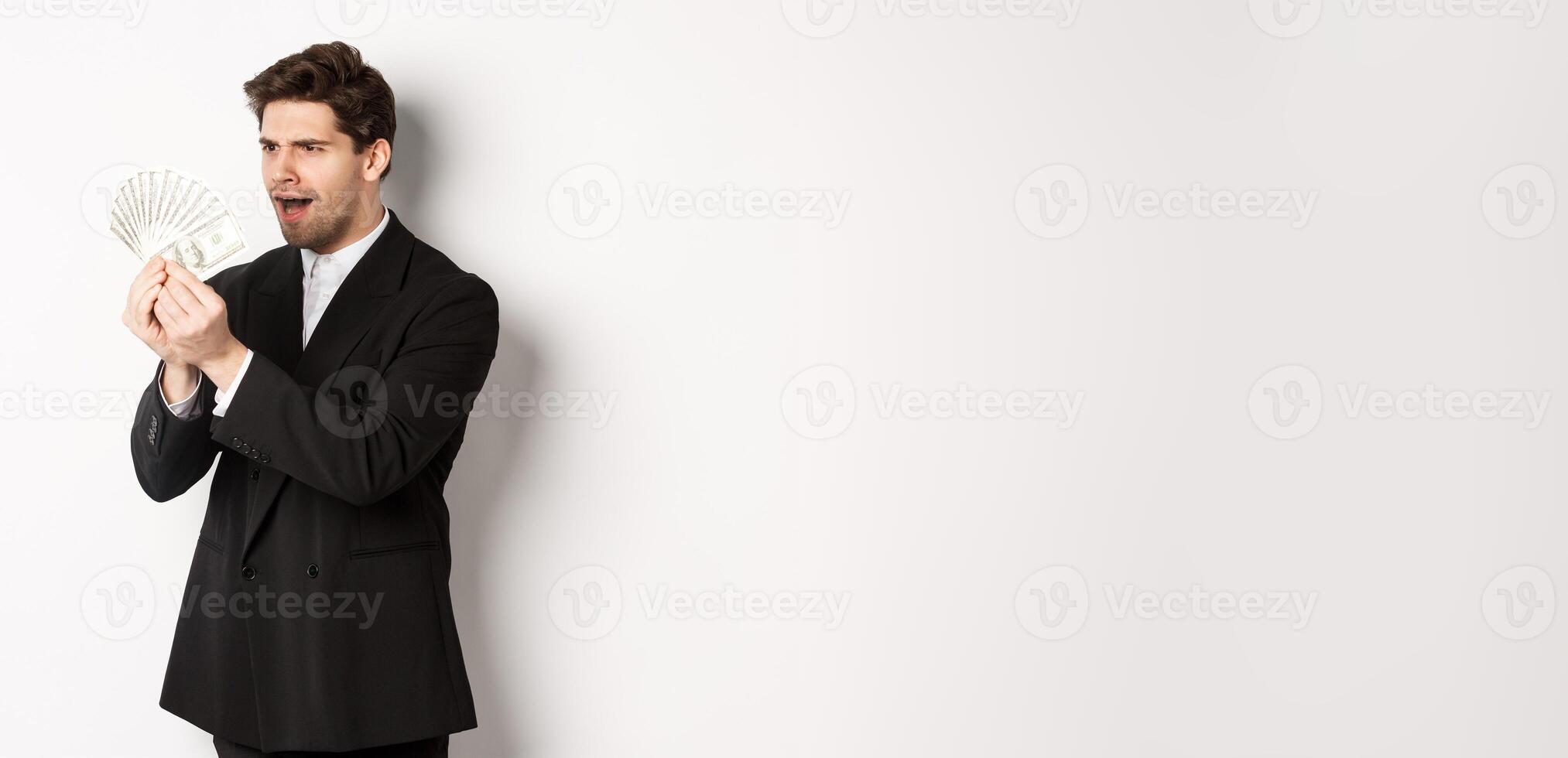 Image of confused businessman looking at fake money, standing over white background in black suit photo