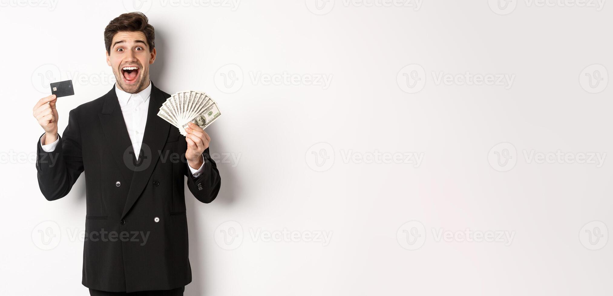Image of handsome male entrepreneur in suit, showing credit card and money, smiling amazed, standing over white background photo