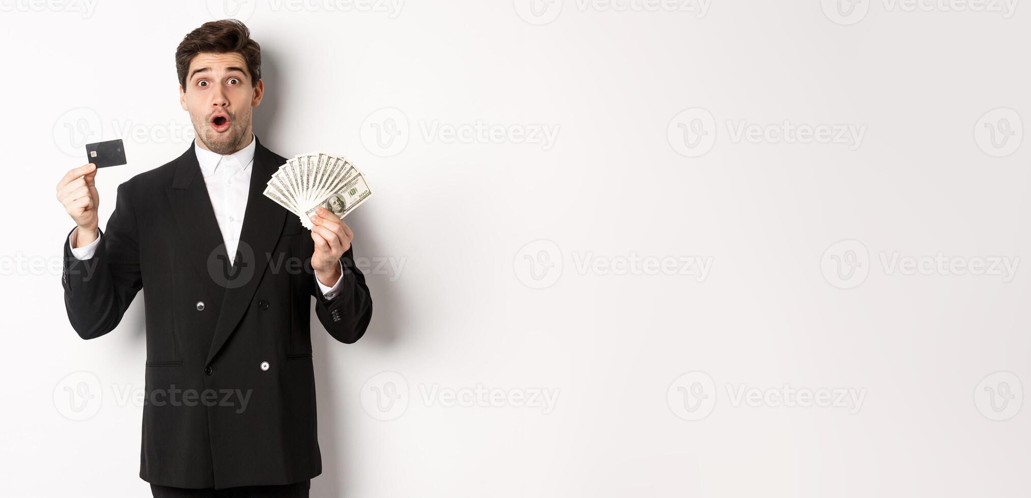 Portrait of surprised handsome man i suit, showing credit card with money, standing against white background photo