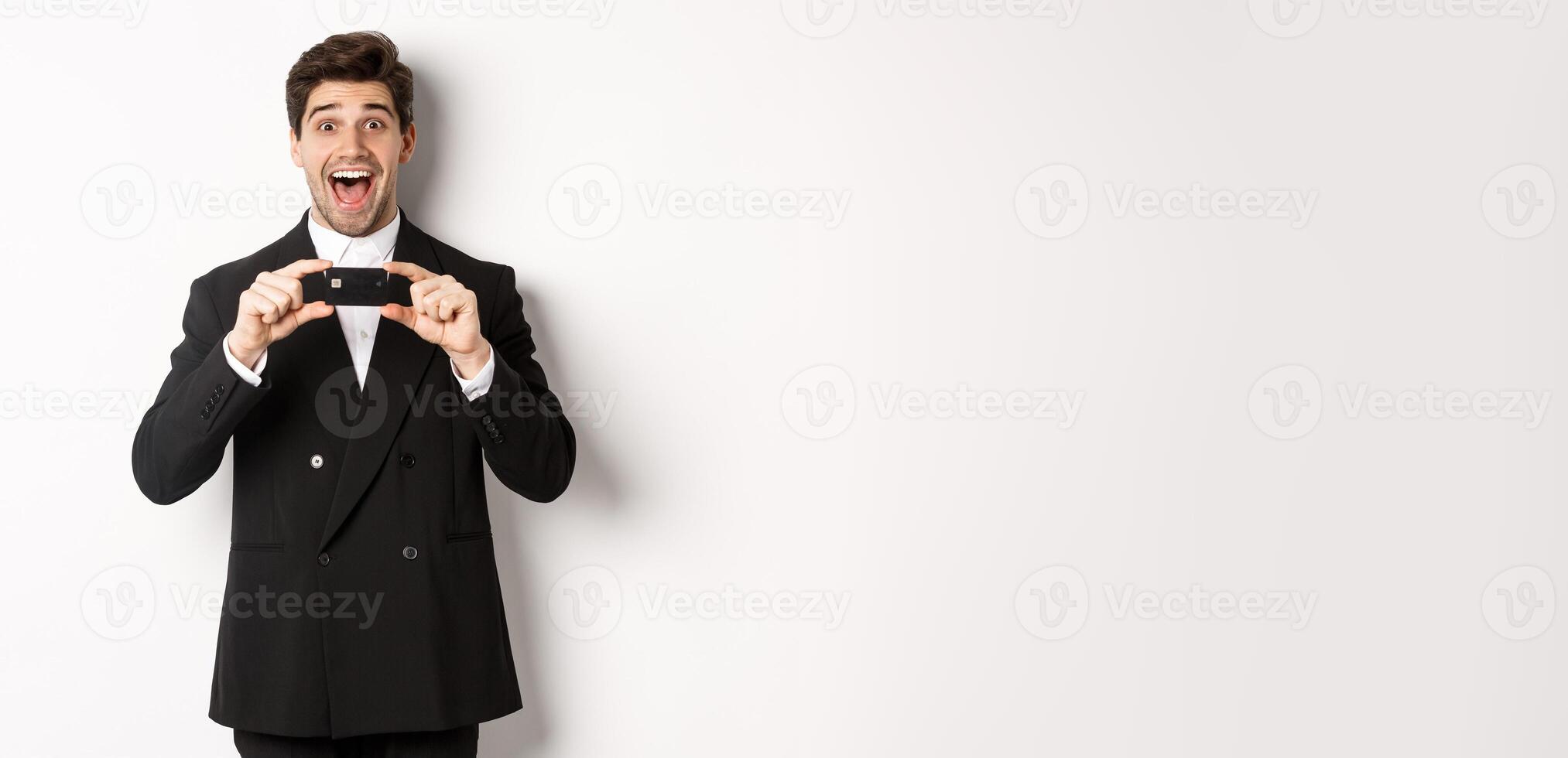 Portrait of amazed handsome man in black suit, showing credit card and recommending bank, standing over white background photo
