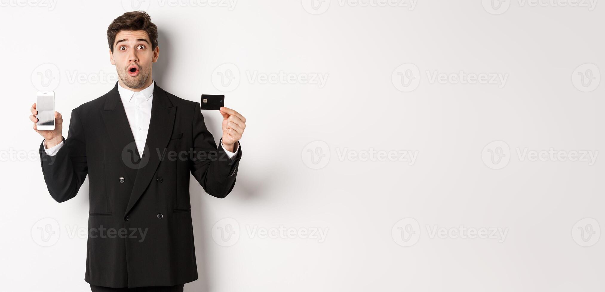 Image of handsome businessman in black suit, looking amazed and showing credit card with mobile phone screen, standing against white background photo