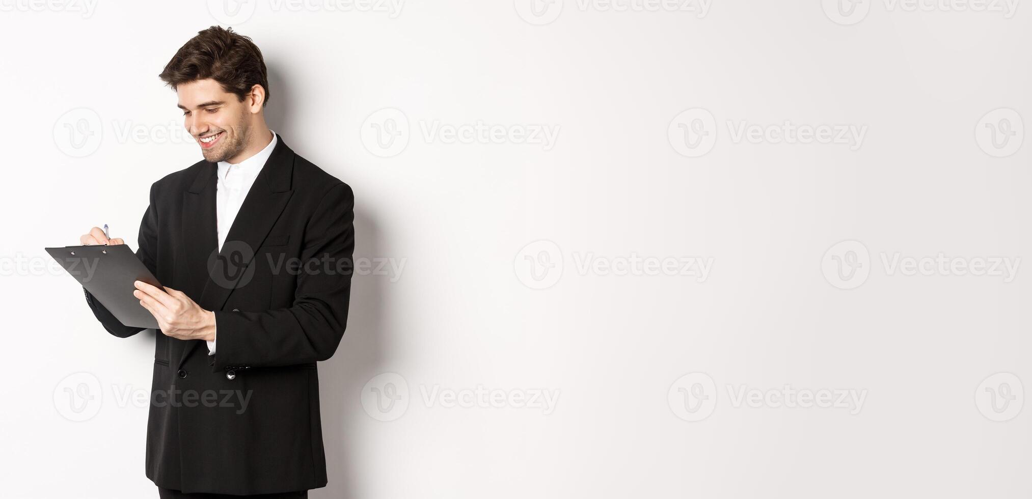 Image of handsome businessman in suit signing documents, looking at clipboard and smiling, standing against white background photo