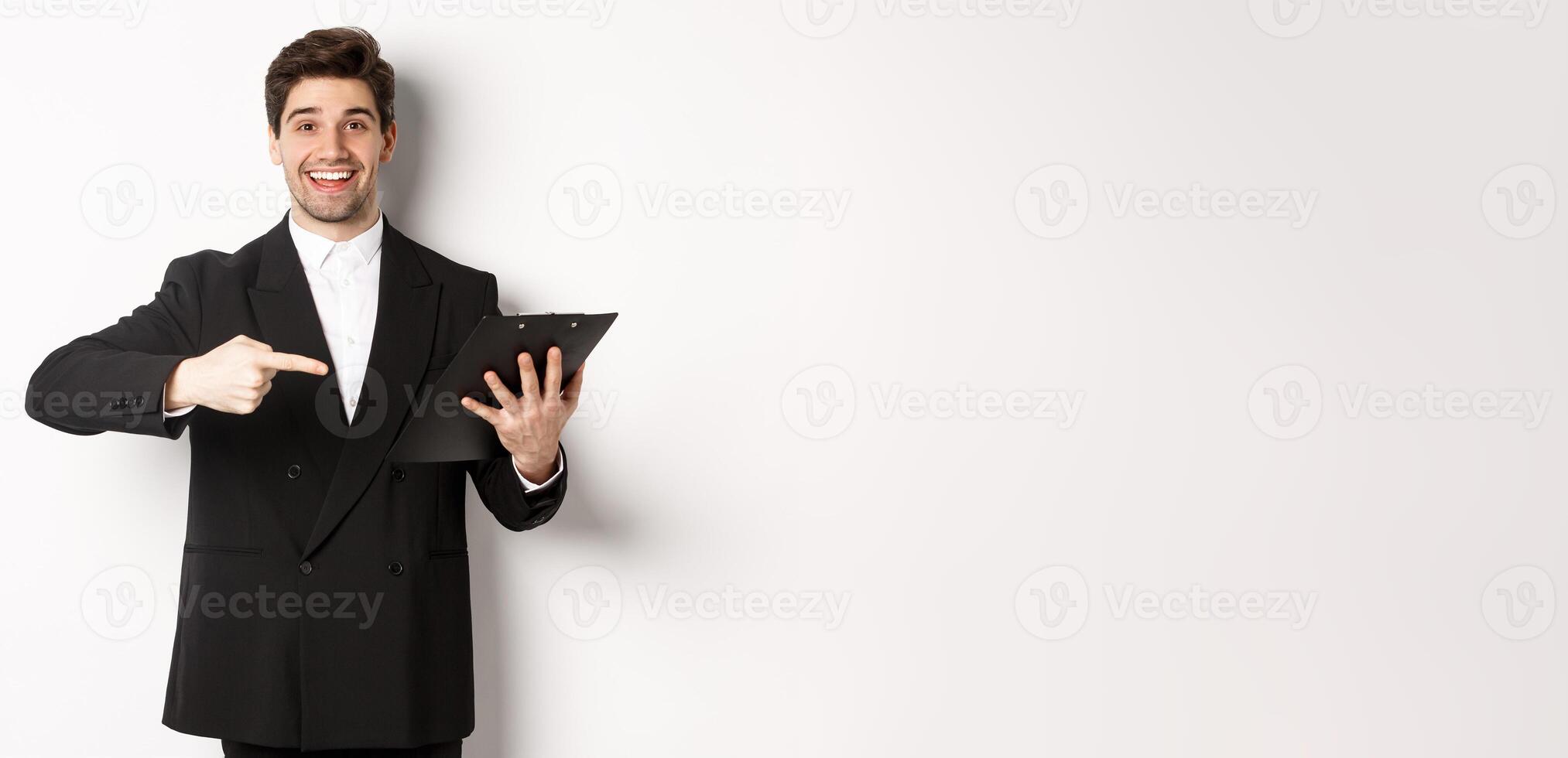 Image of handsome smiling businessman in black suit, pointing finger at clipboard with documents, standing against white background photo