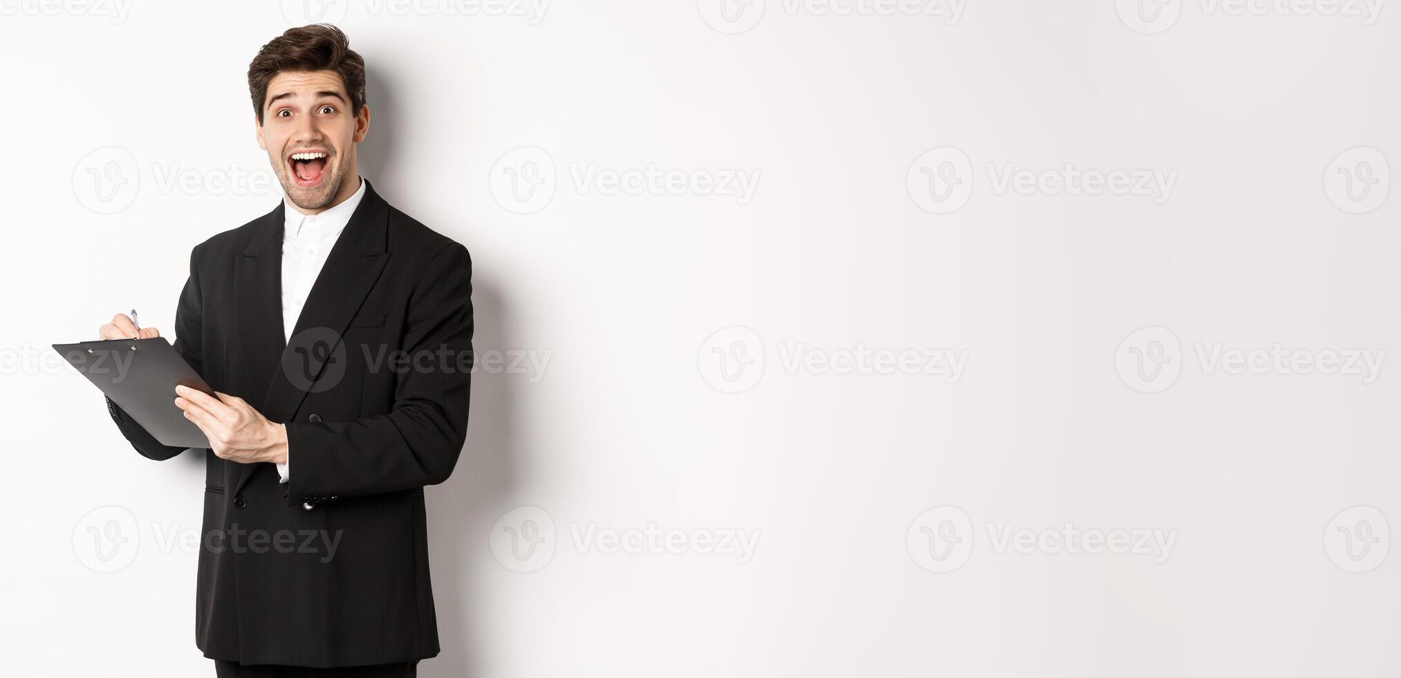 Portrait of handsome businessman in black suit, looking amazed while signing documents on clipboard, standing against white background photo