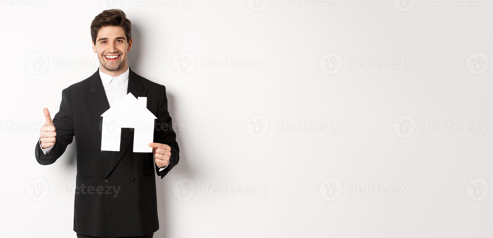Portrait of confident real estate agent showing house maket and thumb-up, standing against white background photo