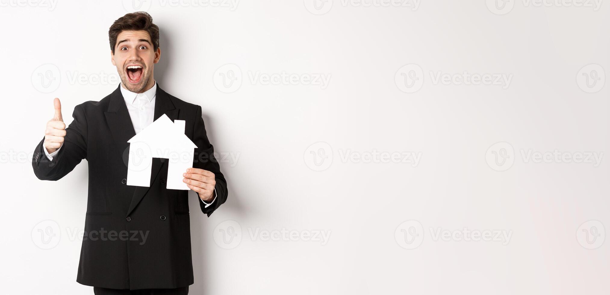 Portrait of excited handsome man looking for a home, showing house and thumb-up, recommending real estate agency, standing against white background photo
