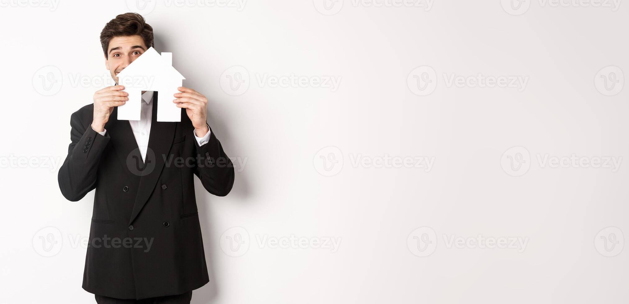 Image of handsome broker in black suit, showing house maket and smiling, selling homes, standing against white background photo