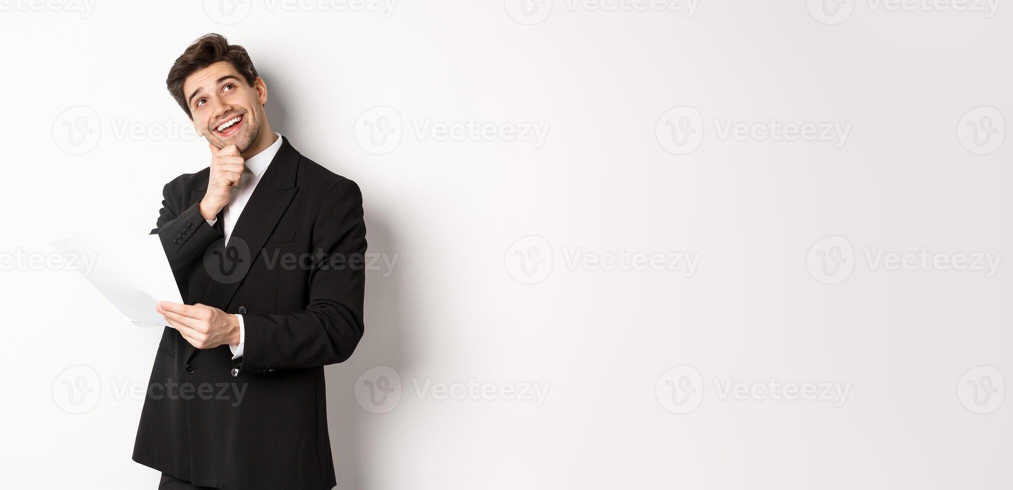 Image of thoughtful, handsome businessman in black suit, holding document and looking at upper right corner, thinking and smiling, standing against white background photo